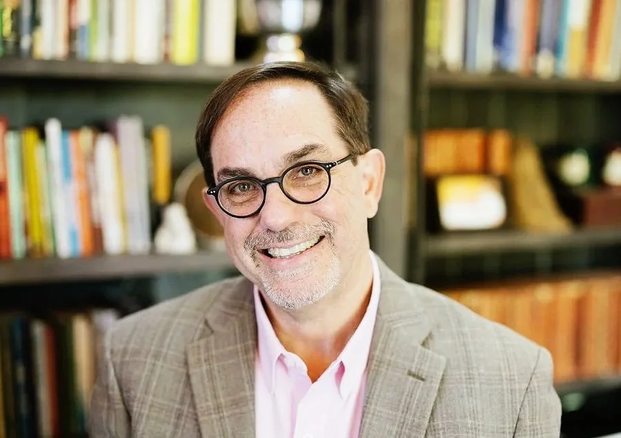 A man with glasses and a beard smiling, wearing a light-colored blazer and pink shirt in front of a bookshelf.