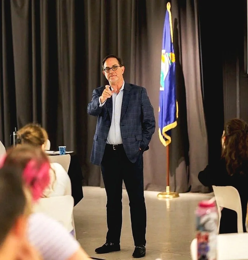 A man in a suit and glasses stands in front of a black curtain, pointing towards the camera, with a flag in the background. People are seated around him in a meeting or seminar setting.