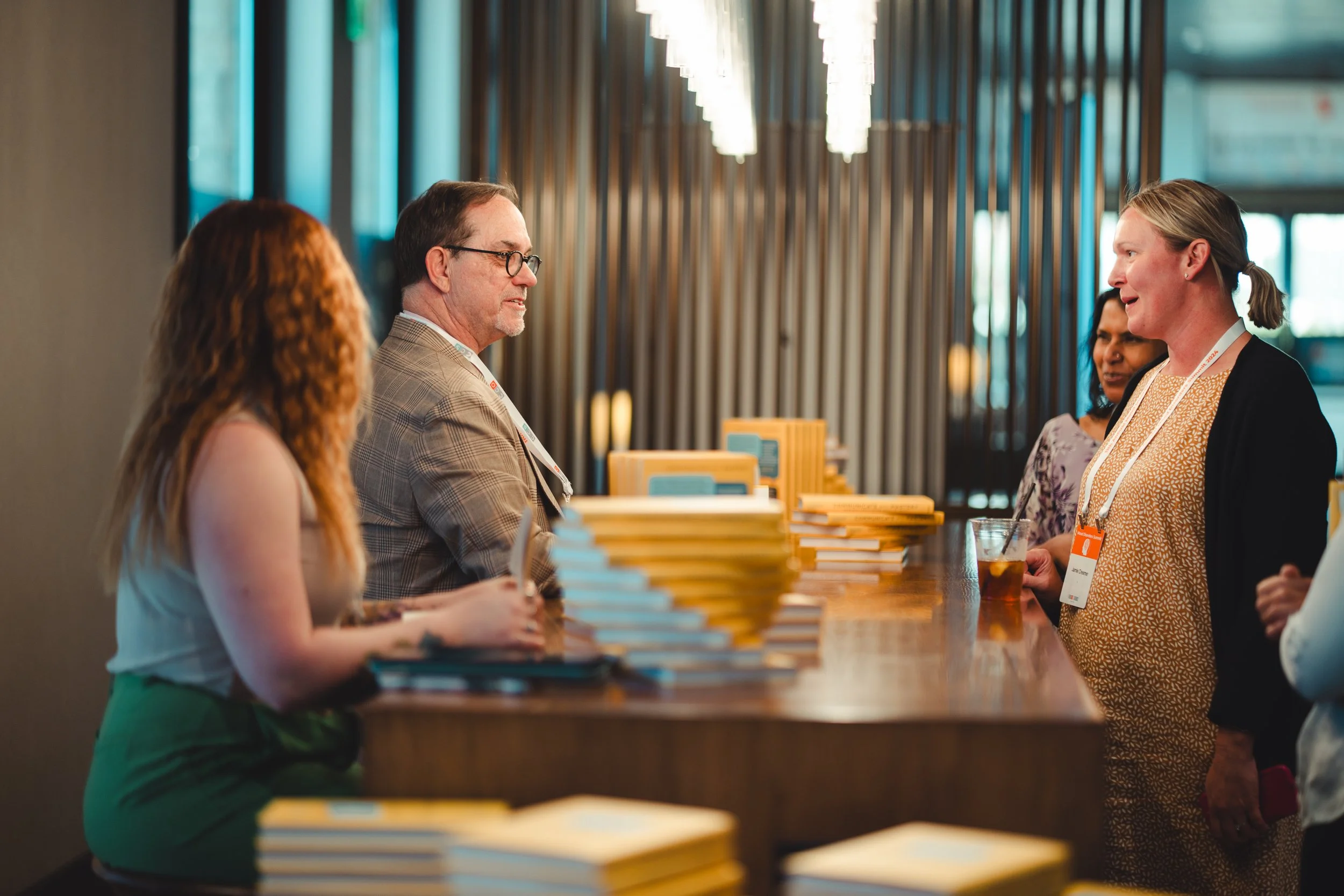 People at a registration or check-in desk at a conference or event, with stacks of yellow books or notebooks on the counter.