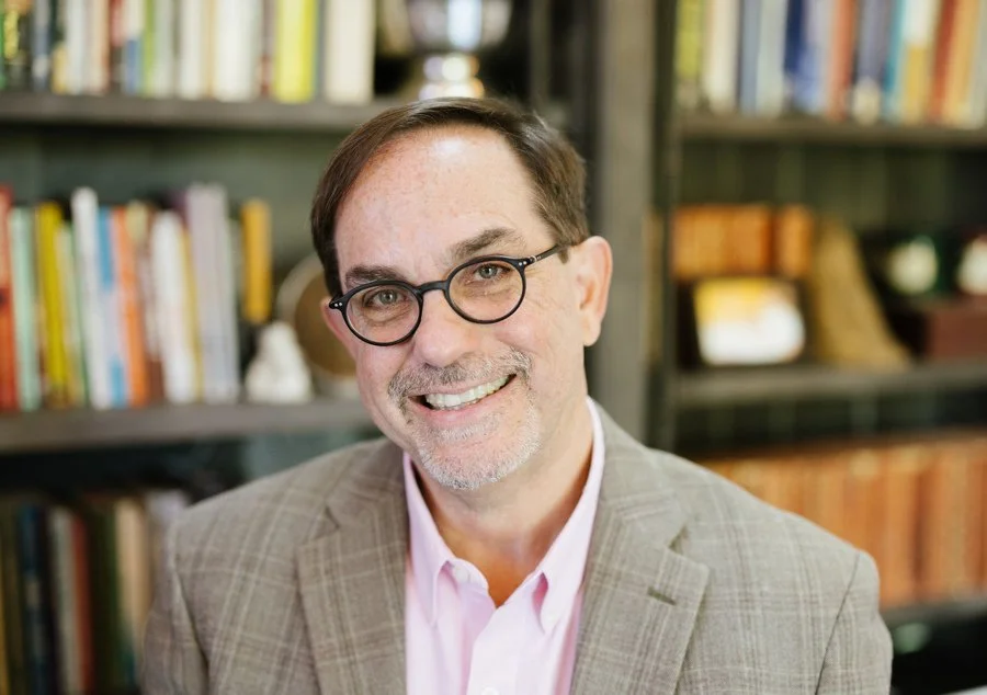 JD Schramm smiling & wearing a light-colored blazer and pink shirt, in front of a bookshelf filled with books.