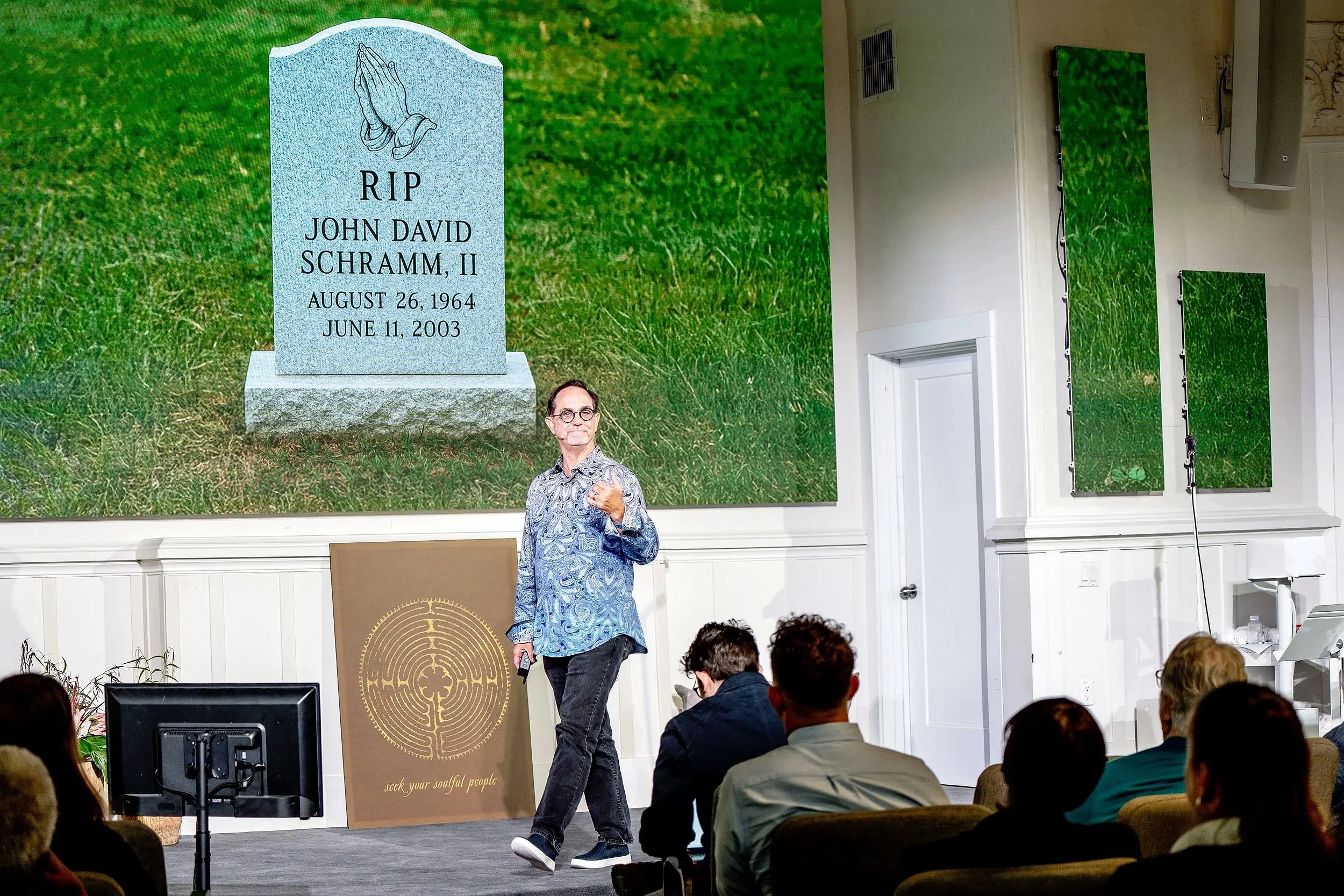 Presenter on stage at a conference, standing in front of a large screen displaying a virtual cemetery gravestone with the name John David Schramm, II, and dates August 26, 1964, to June 11, 2003, in front of a green grass background. Audience members
