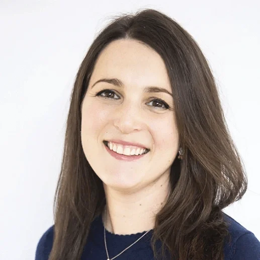 A young woman with long brown hair smiling, wearing earrings and a blue top, against a white background.