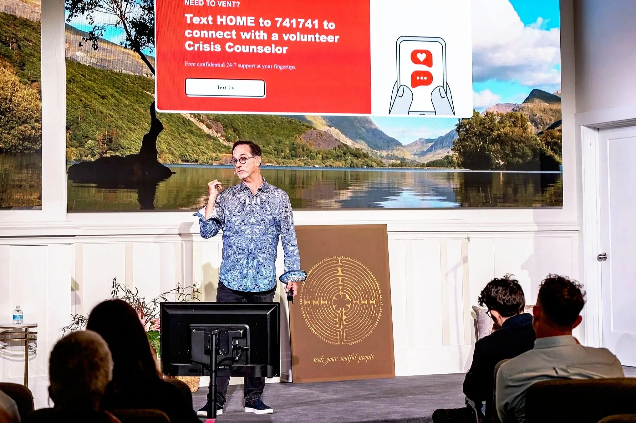 A man giving a presentation in a room with a large landscape mural behind him. There is a large screen above him displaying a red message about connecting with a crisis counselor via text. Audience members are seated in front.