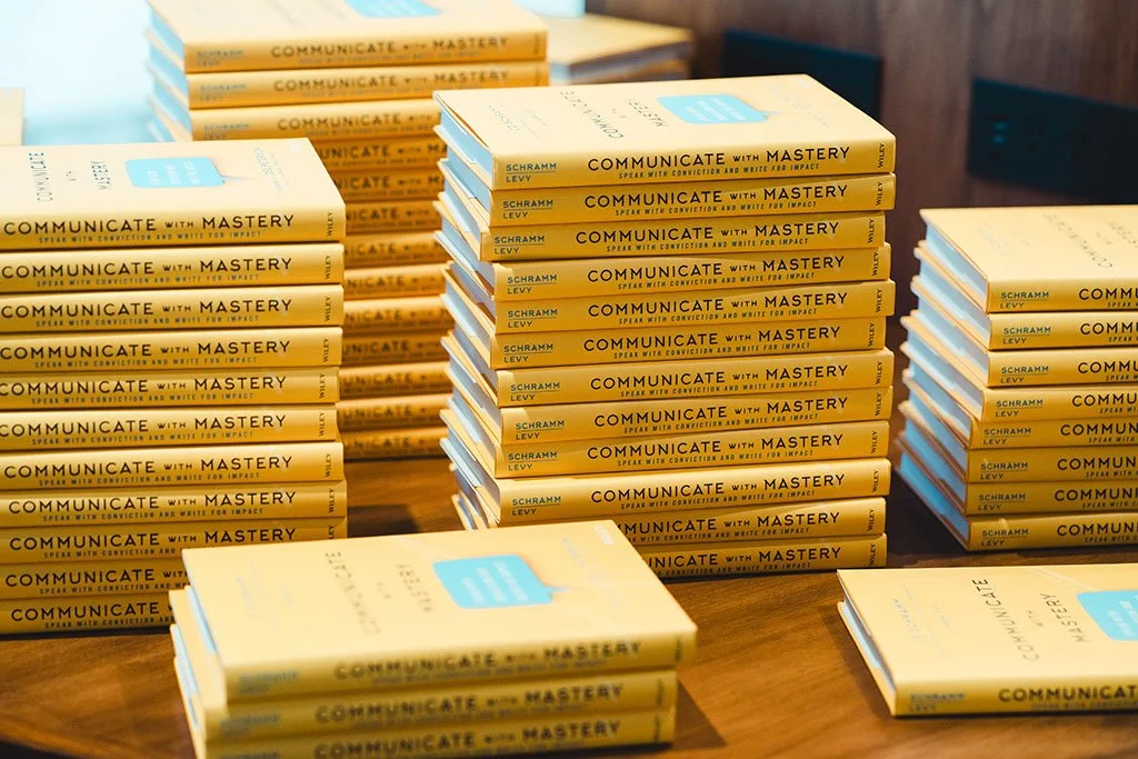Stacks of books titled 'Communicate with Mastery' by Schramm Levy arranged on a wooden table.