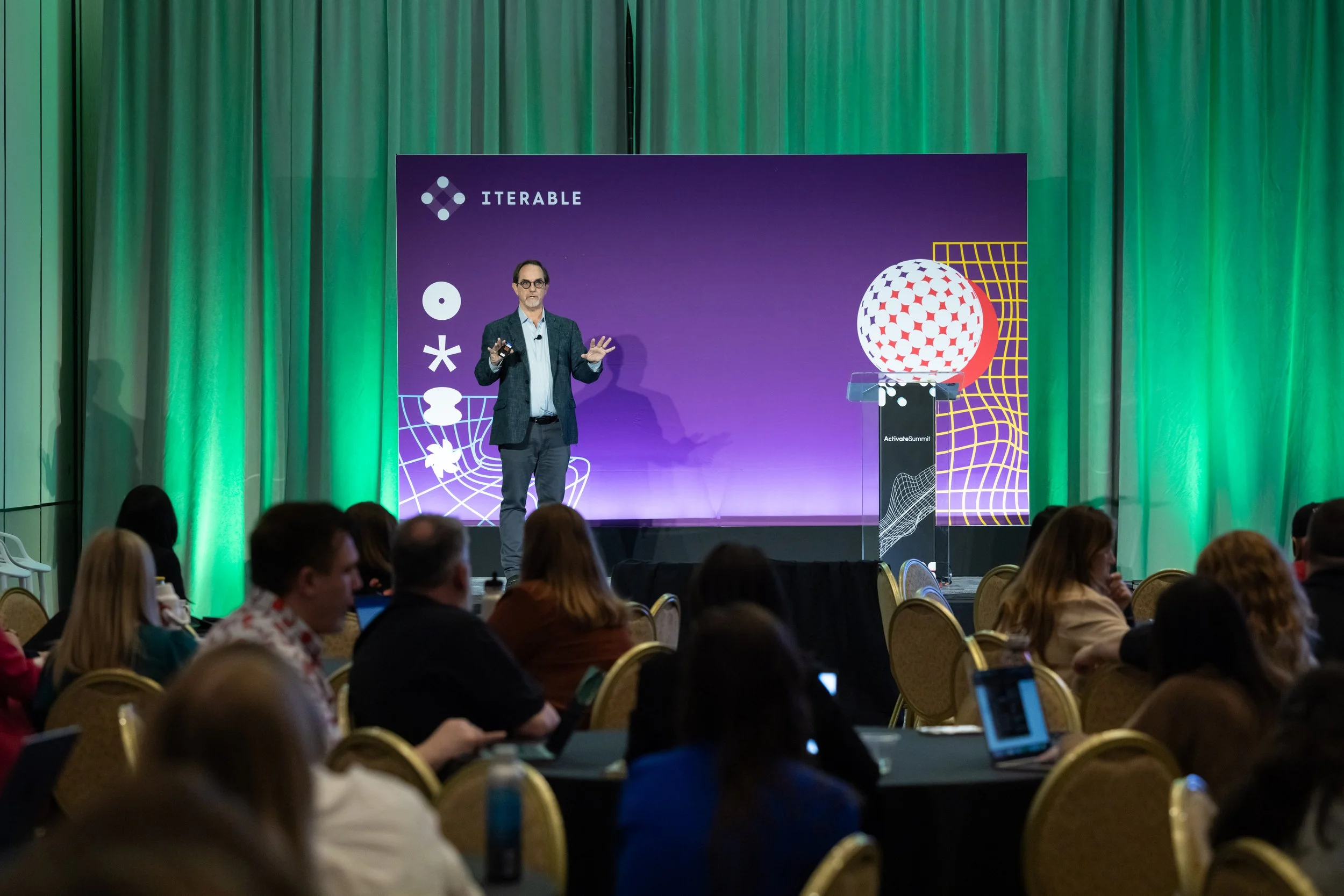 A man giving a presentation on stage at a conference, with a large purple and green backdrop, and an audience seated at round tables.