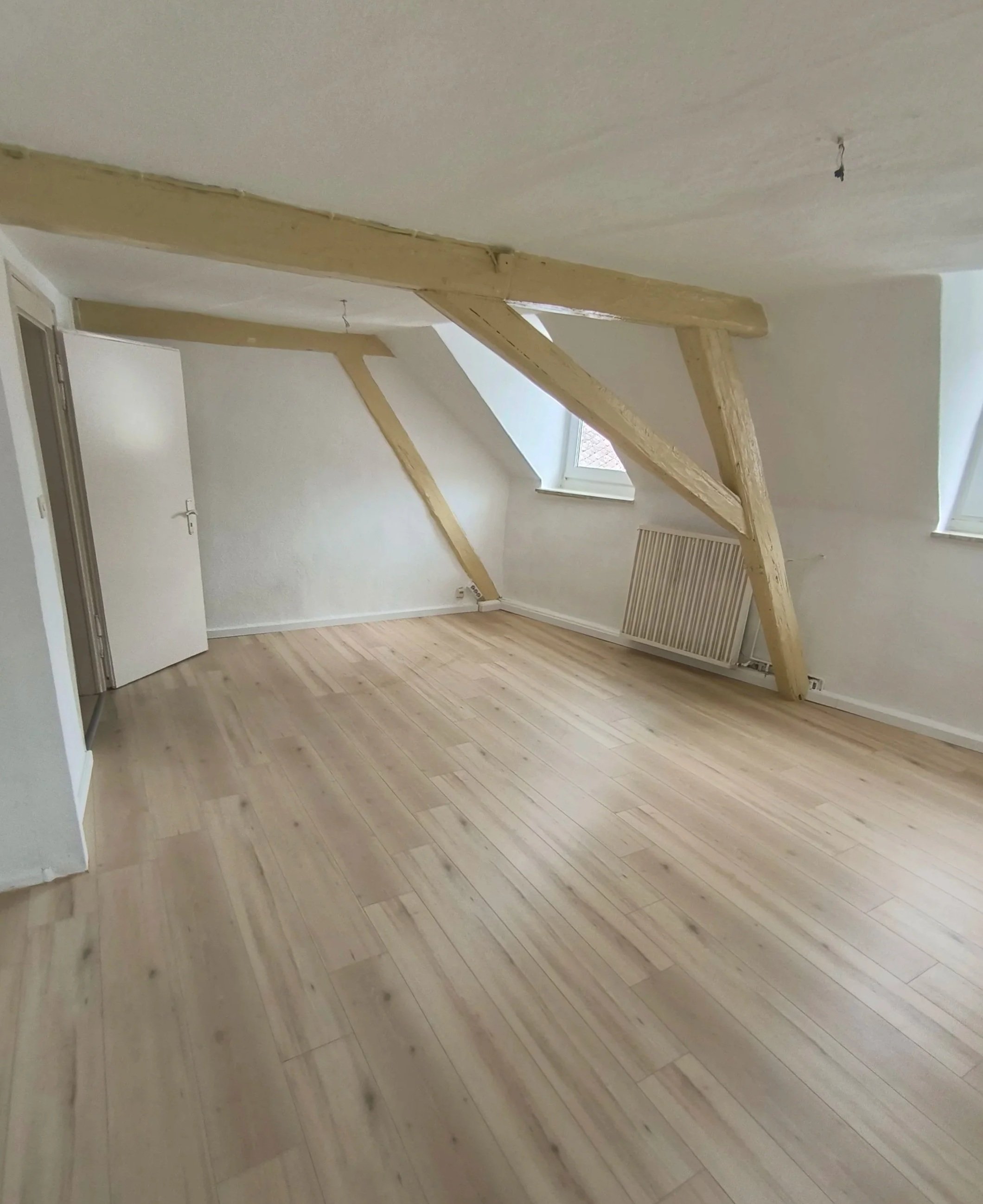 Leeres Zimmer mit Holzbalken an der Decke, zwei Dachfenster und Holzboden.