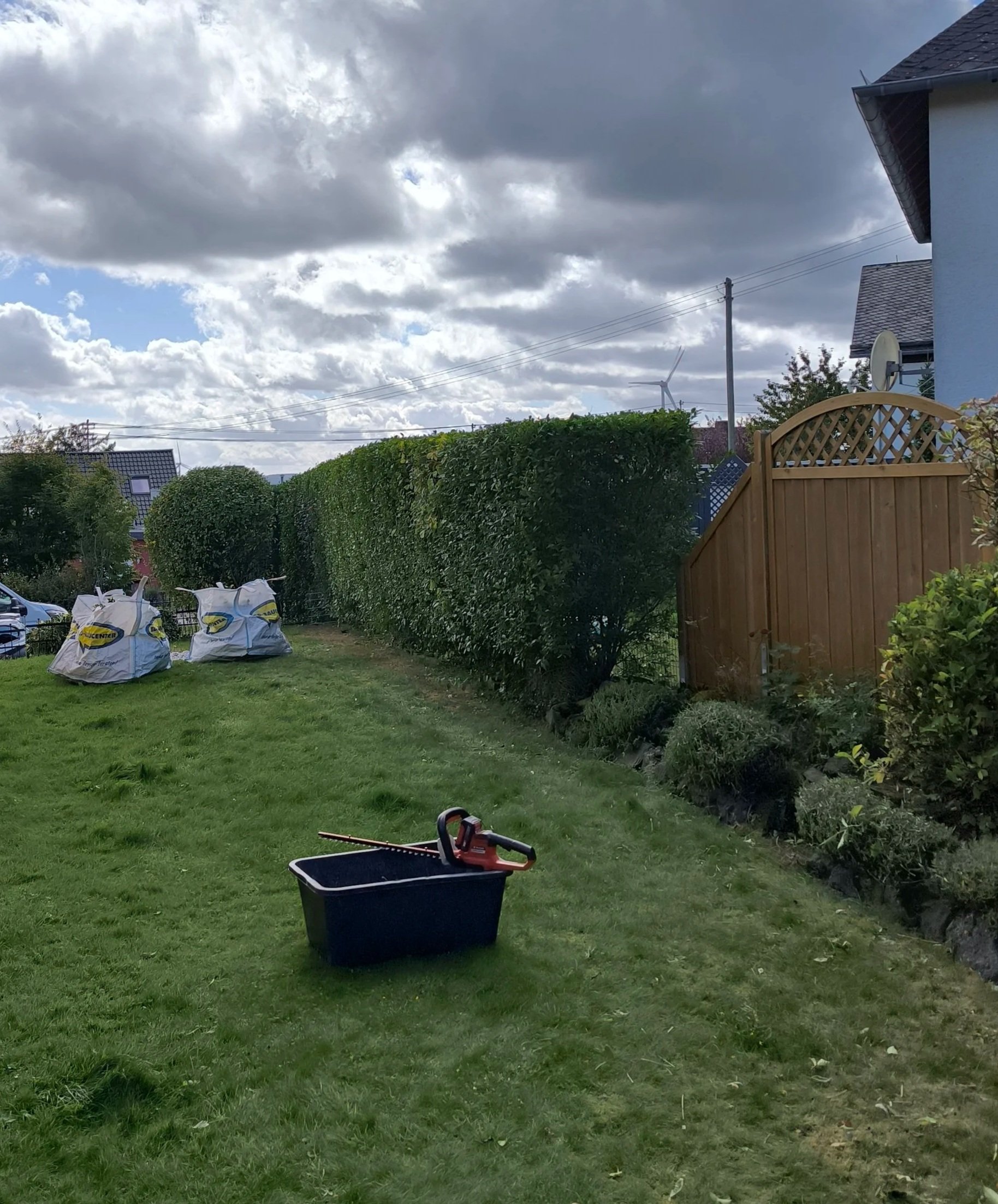 Ein Garten mit grüner Rasenfläche, einem Holzzaun, Büschen und einem Himmel mit Wolken. Im Vordergrund liegt eine schwarze Kiste mit einem Elektrosägen. Im Hintergrund sind drei größe Säcke mit Gartenabfällen.