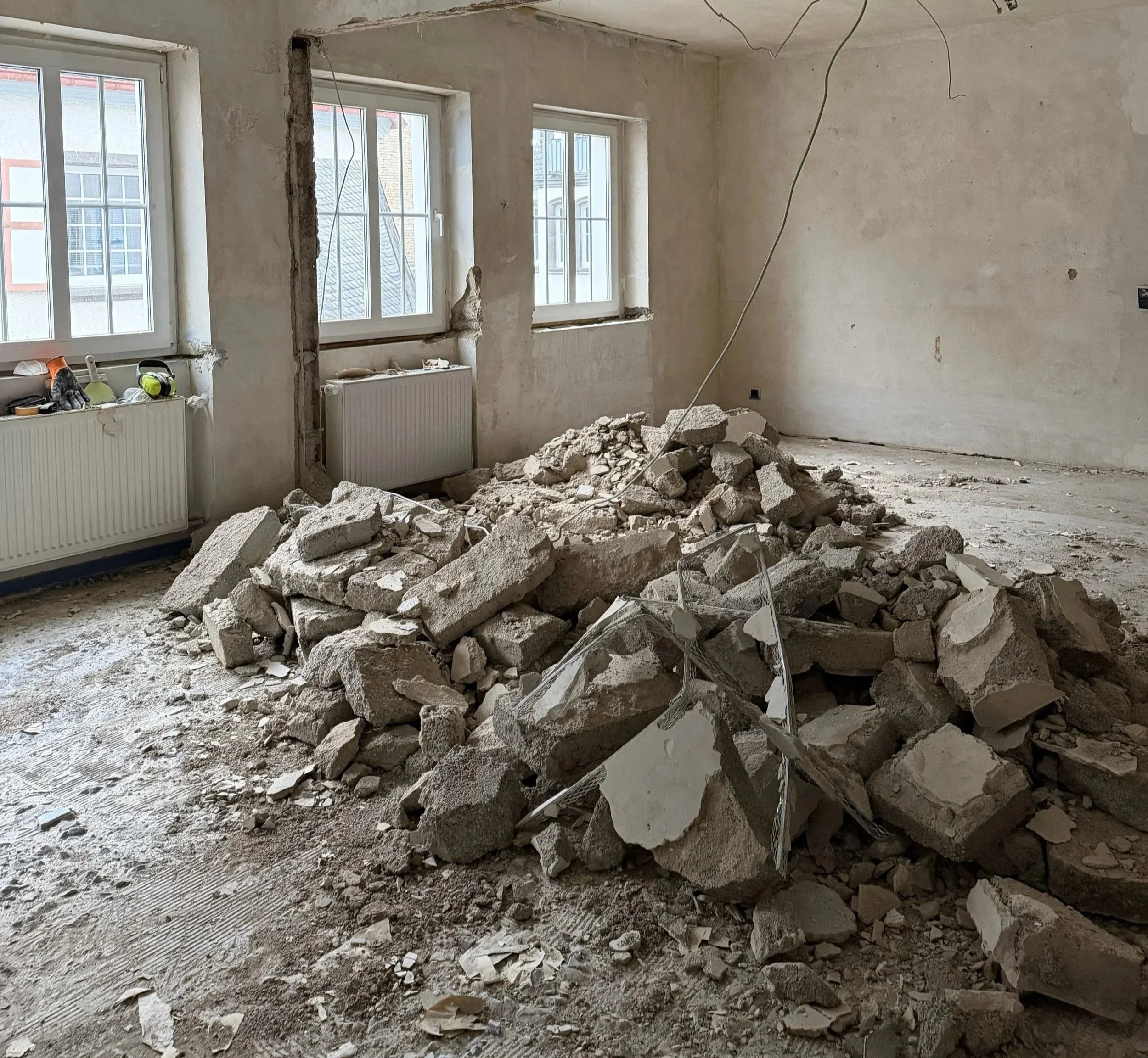 Baufälliger Raum mit zerbrochenen Wänden und einem Haufen Trümmer, Fenster und Heizkörper sichtbar.