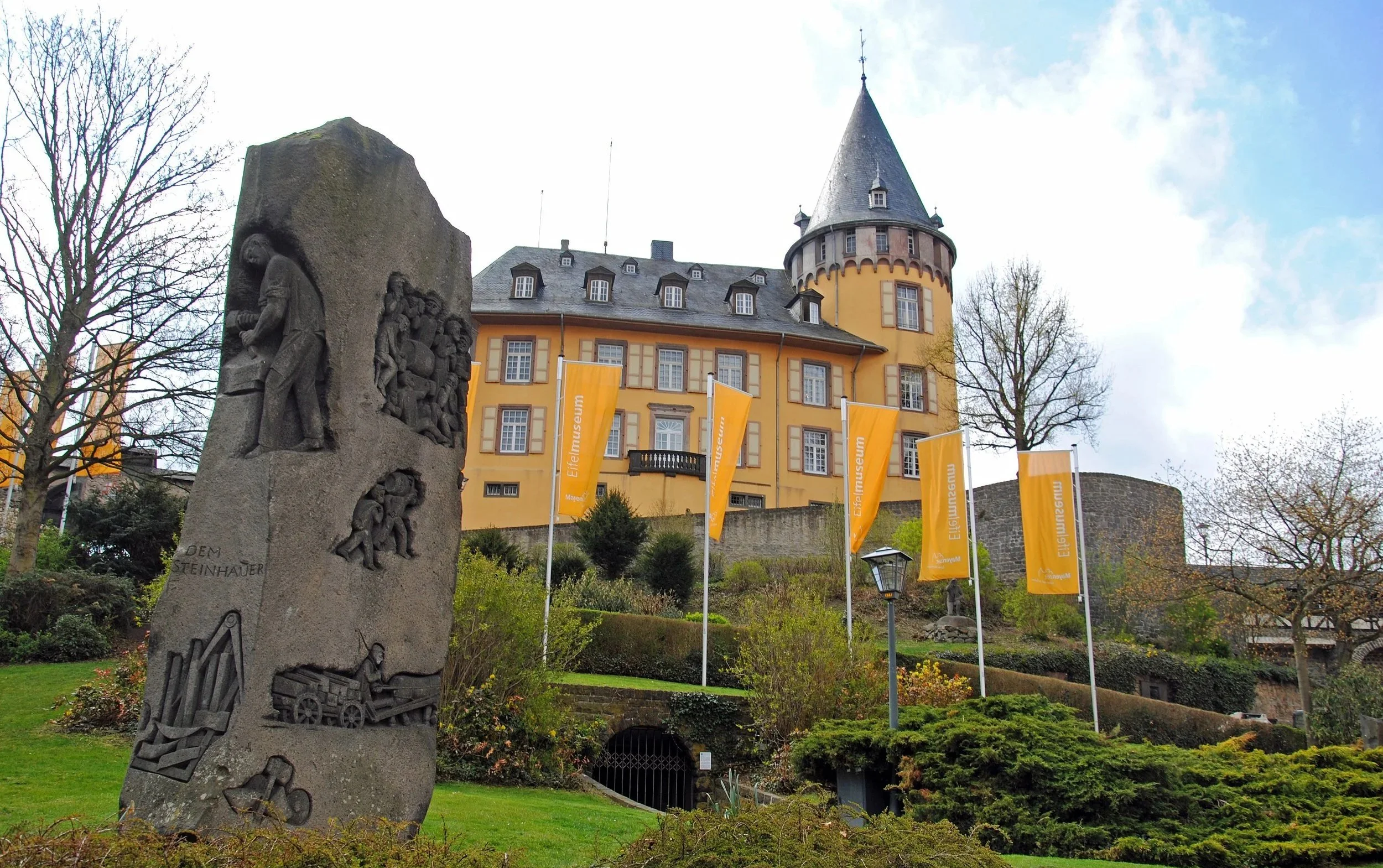 Burg in Mayen mit gelber Fassade und turmartigem Dach, umgeben von Bäumen und Flaggen mit der Aufschrift 'Eisenmuseum' auf einem Hügel.