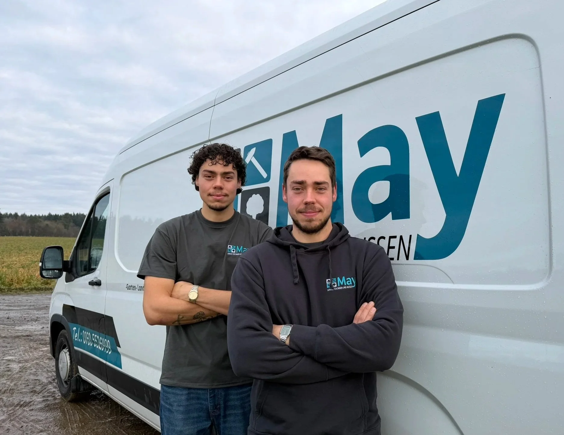 Zwei Männer stehen mit verschränkten Armen vor einem weißen Transporter mit einem großen Firmenlogo und Kontaktinformationen auf einer ländlichen Straße bei bewölktem Himmel.