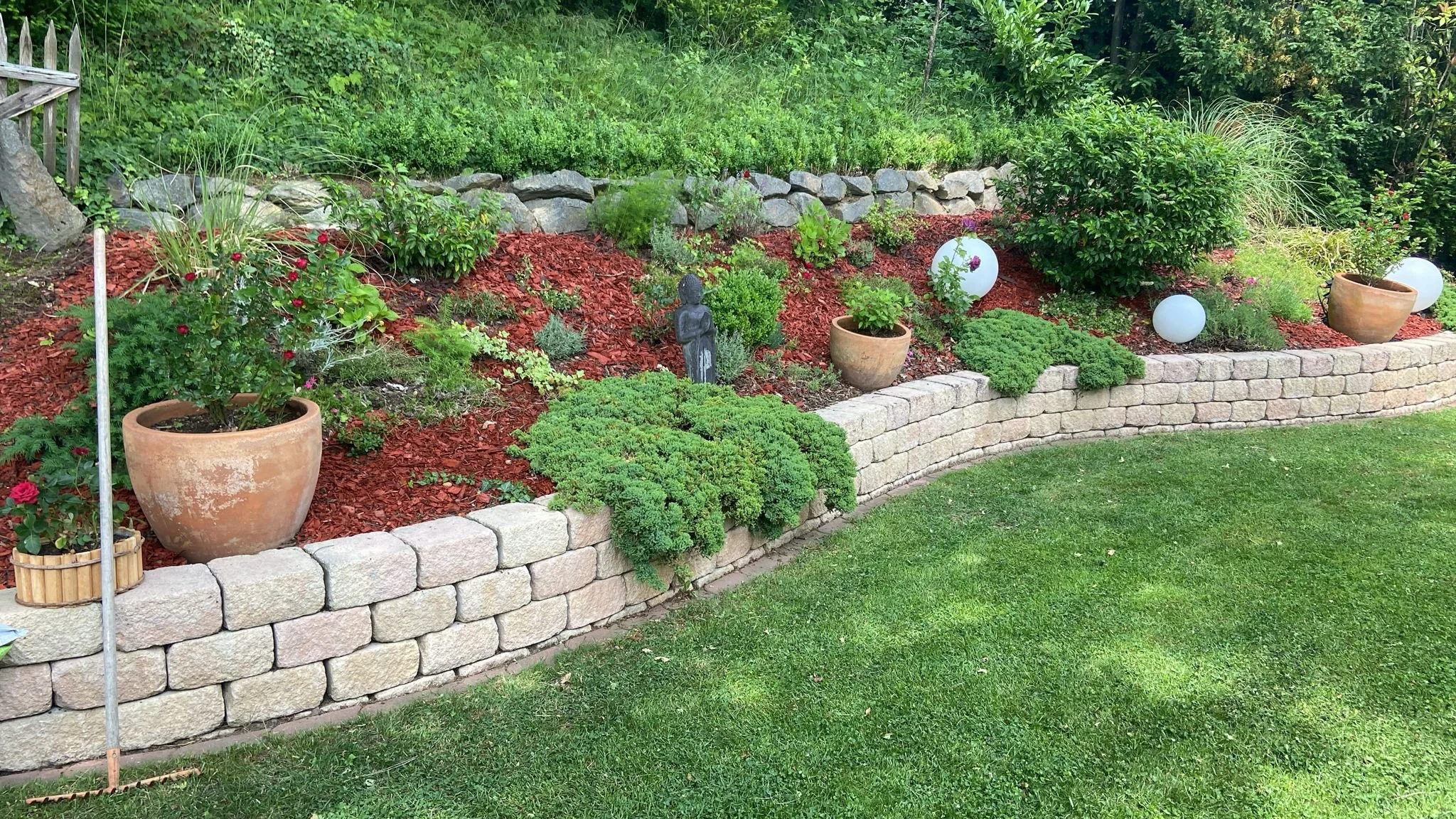 Garten mit Steinmauer, verschiedene Pflanzen in Töpfen und Dekorationen, inklusive runden weißen Lampen, auf einem Rasen