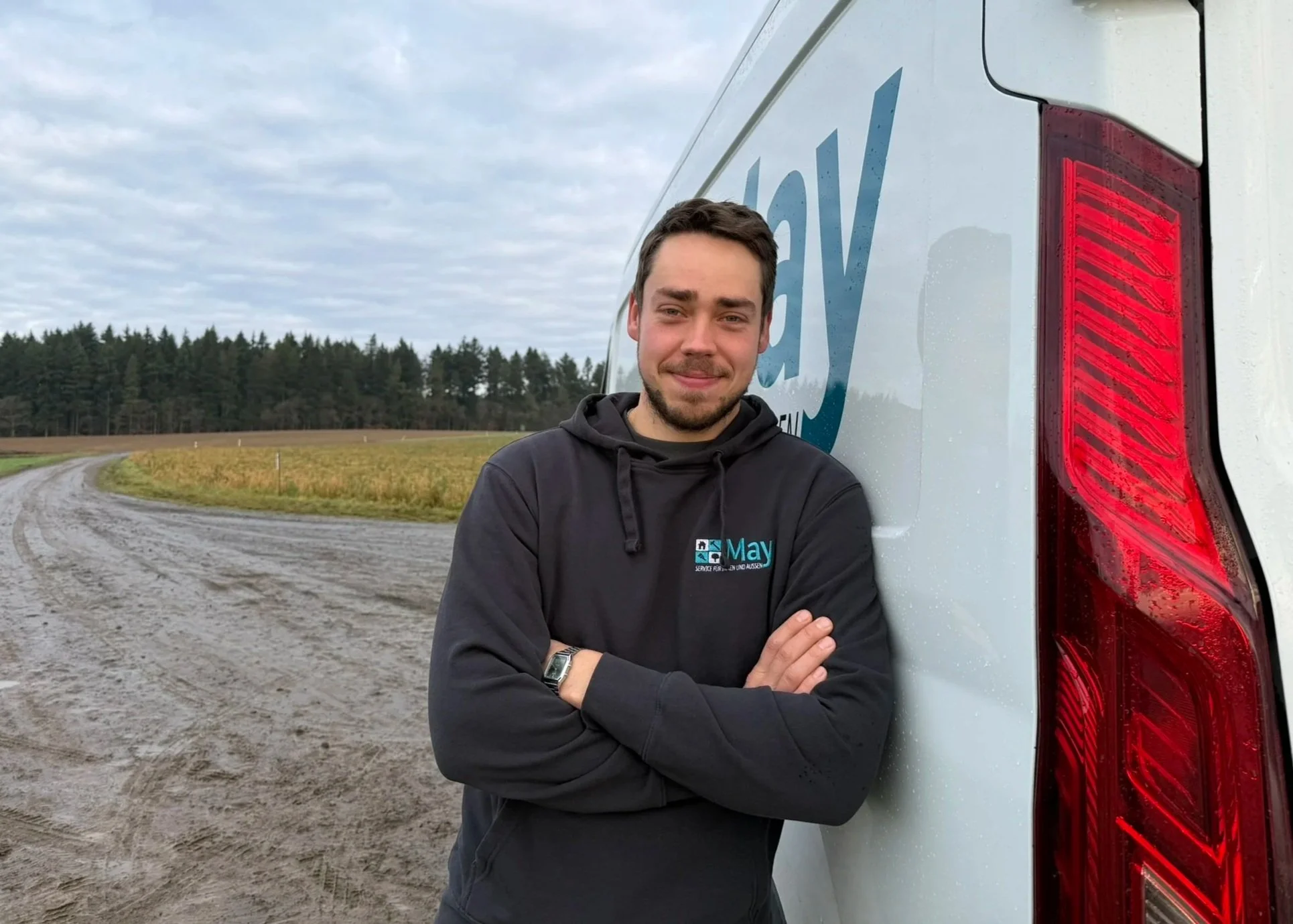 Ein junger Mann mit dunklem Hoodie steht an einer landwirtschaftlichen Straße neben einem weißen Lieferwagen mit Firmenlogo, der im Freien bei bewölktem Himmel steht.