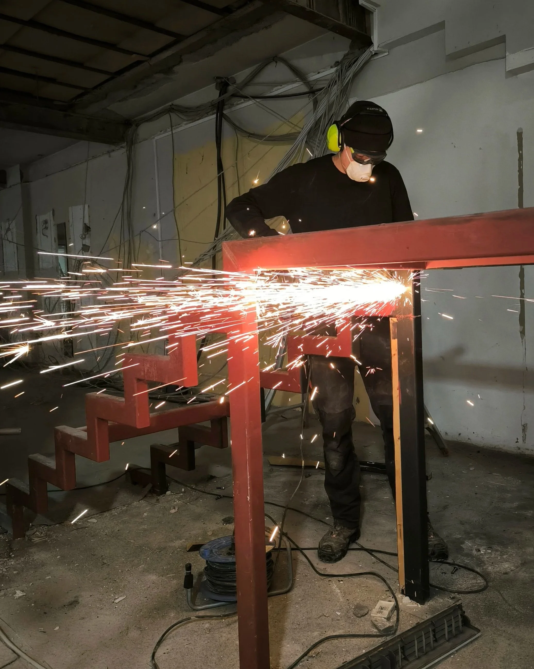 Person entfernt eine rote Metalltreppe. Es sprühen Funken, Person trägt Schutzbrille, Gehörschutz und Maske, im Hintergrund Kabel und Baumaterialien in einem Bauwerk.