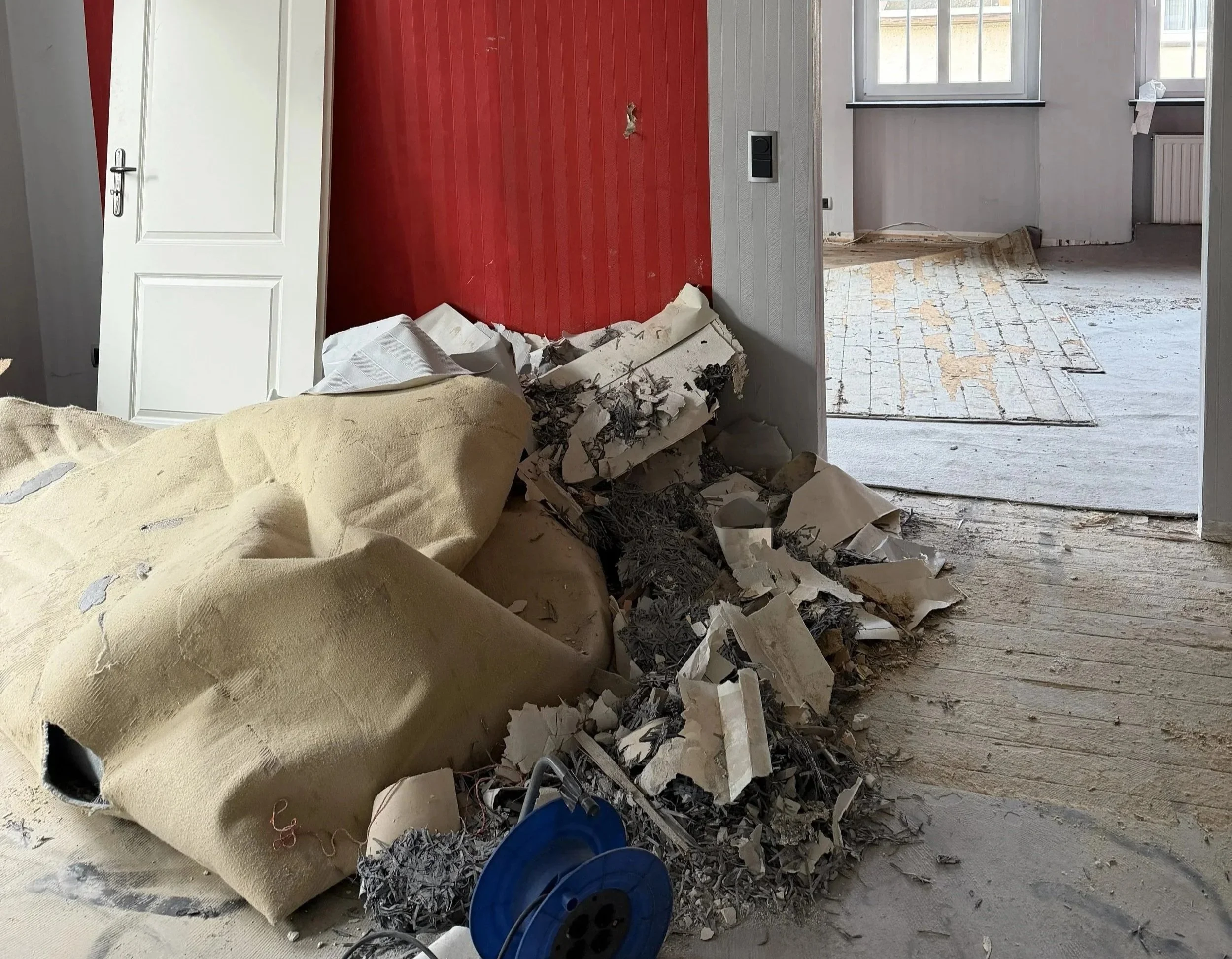 Renovierungsarbeiten in einem Raum mit entferntem Teppich und Müll auf dem Boden, offene Türen und Fenster, die Licht in den Raum lassen.