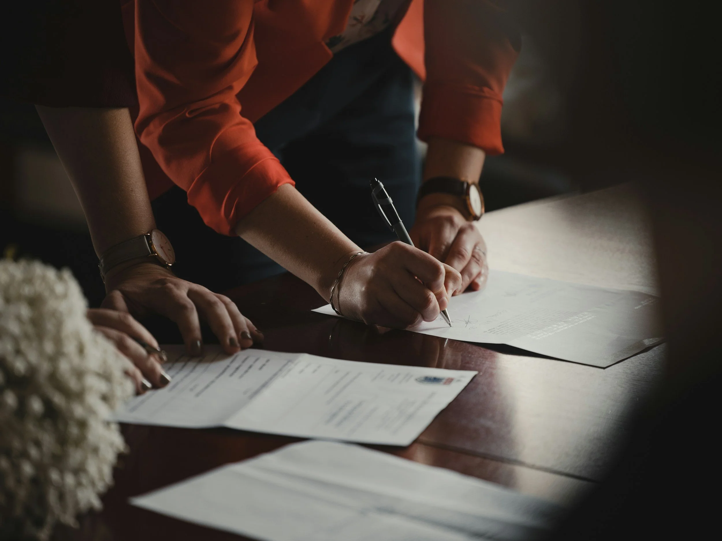 People signing documents on a wooden table with scattered papers and flowers.
