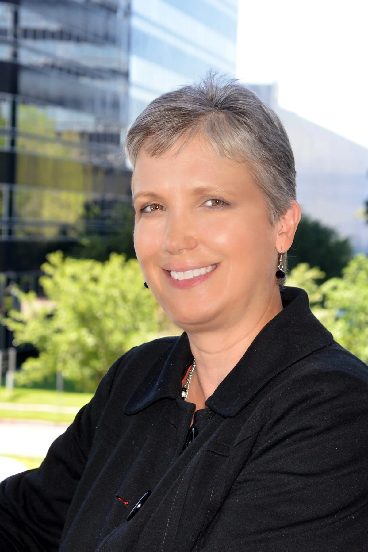 A woman with short gray hair smiling outdoors, wearing a black coat and earrings, with a modern glass building and trees in the background.