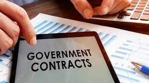 Tablet screen displaying the text 'Government Contracts', with a person's hands typing on a keyboard nearby and financial charts in the background.