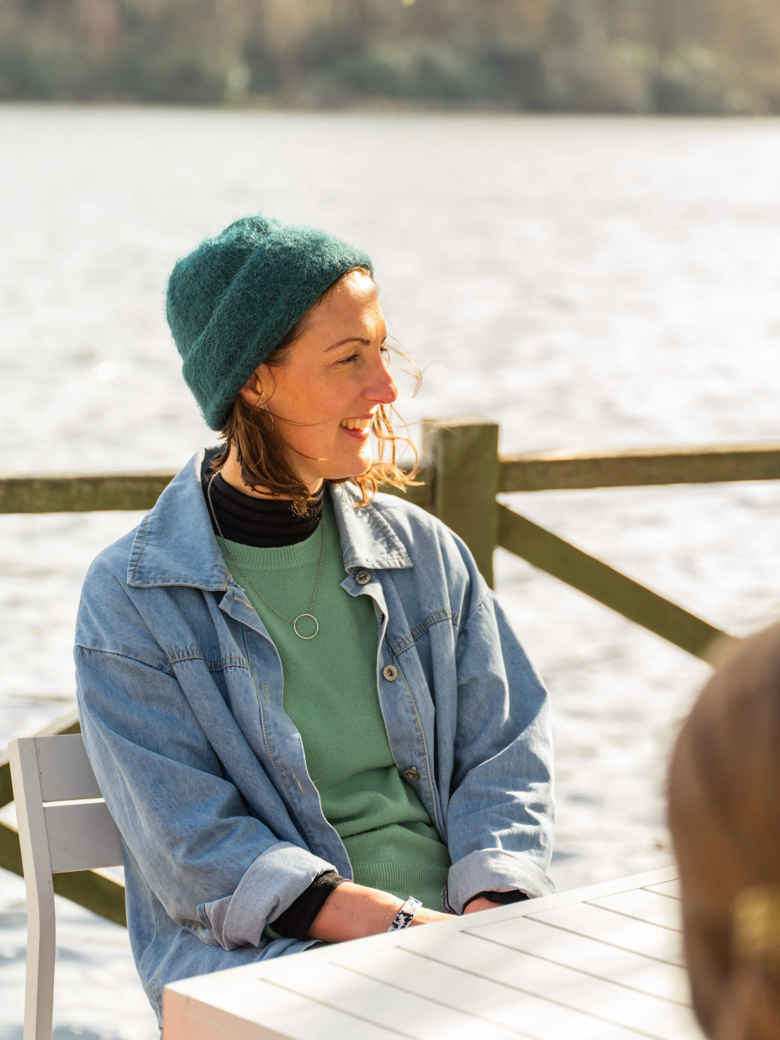 A woman wearing a green beanie and denim jacket sitting outdoors by a body of water, smiling and talking to someone off-camera.