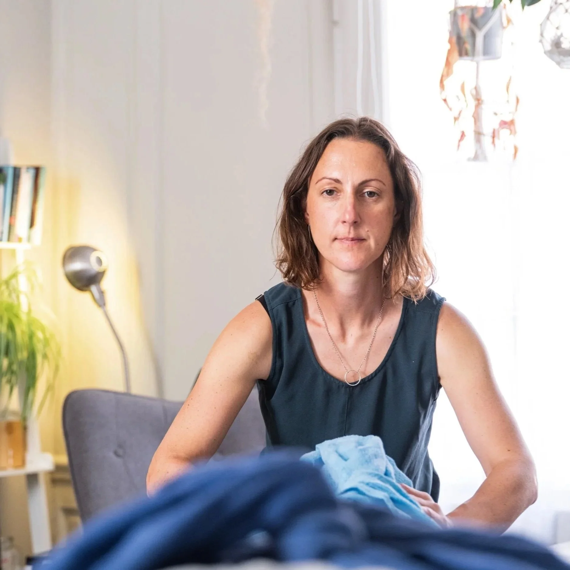 A woman with shoulder-length brown hair, wearing a navy sleeveless top, sitting indoors near a window with natural light, facing the camera with a neutral expression. In the background, there is a lamp, a bookshelf, and a houseplant.