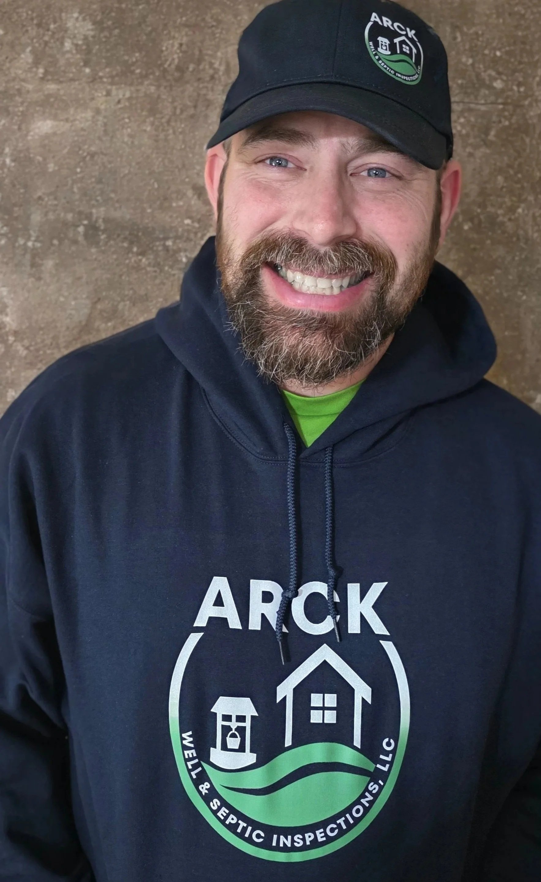 A man wearing a black cap and hoodie with the logo of ARCK Well & Septic Inspections, LLC, smiling, with a brown textured background.