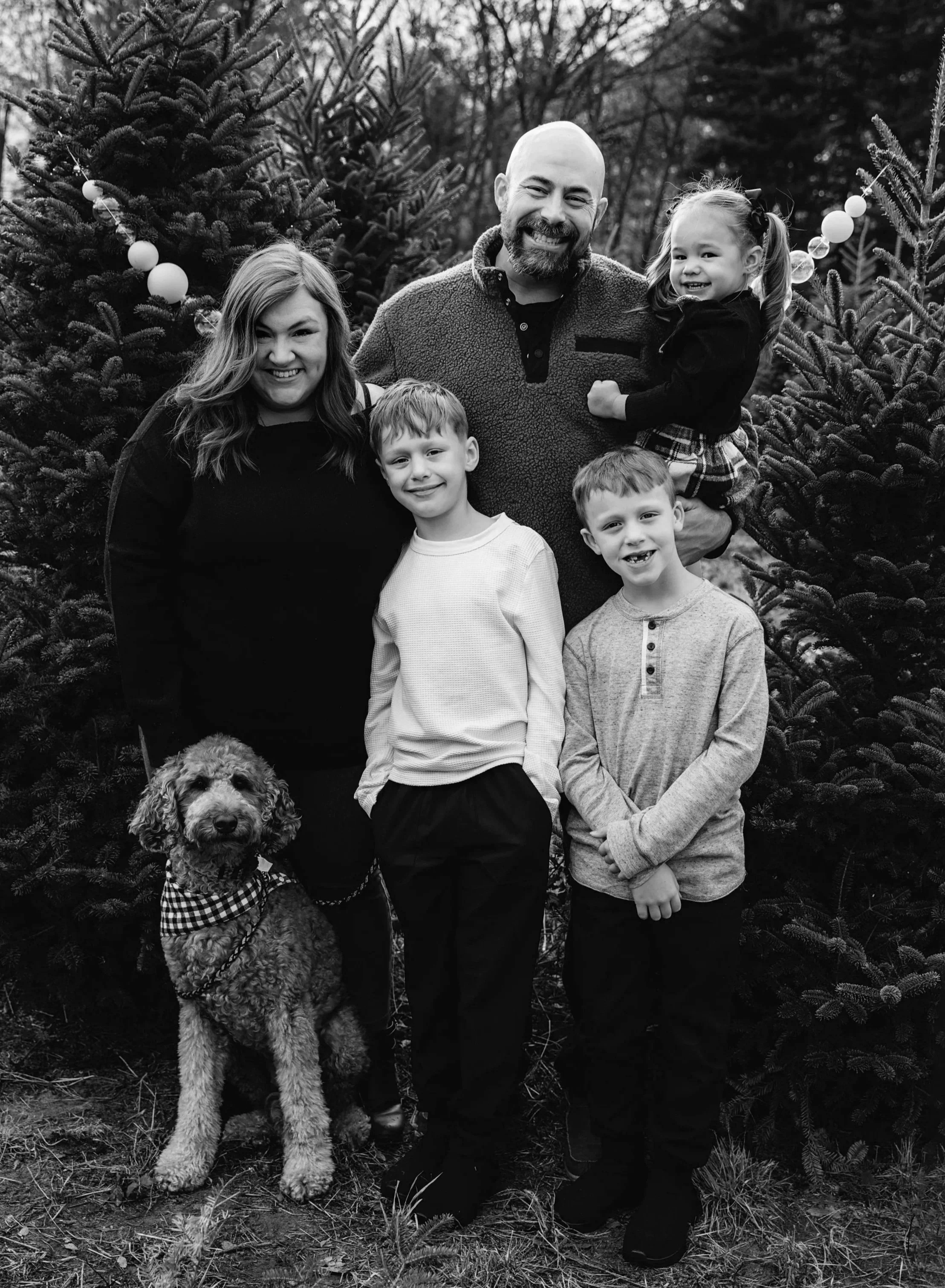 A family of six, including a woman, a man, three children, and a dog, standing outdoors in front of Christmas trees. The woman and children have light hair, and the man has a bald head and beard. One girl is being carried by the man, and the dog is sitting at the woman's side, all smiling.