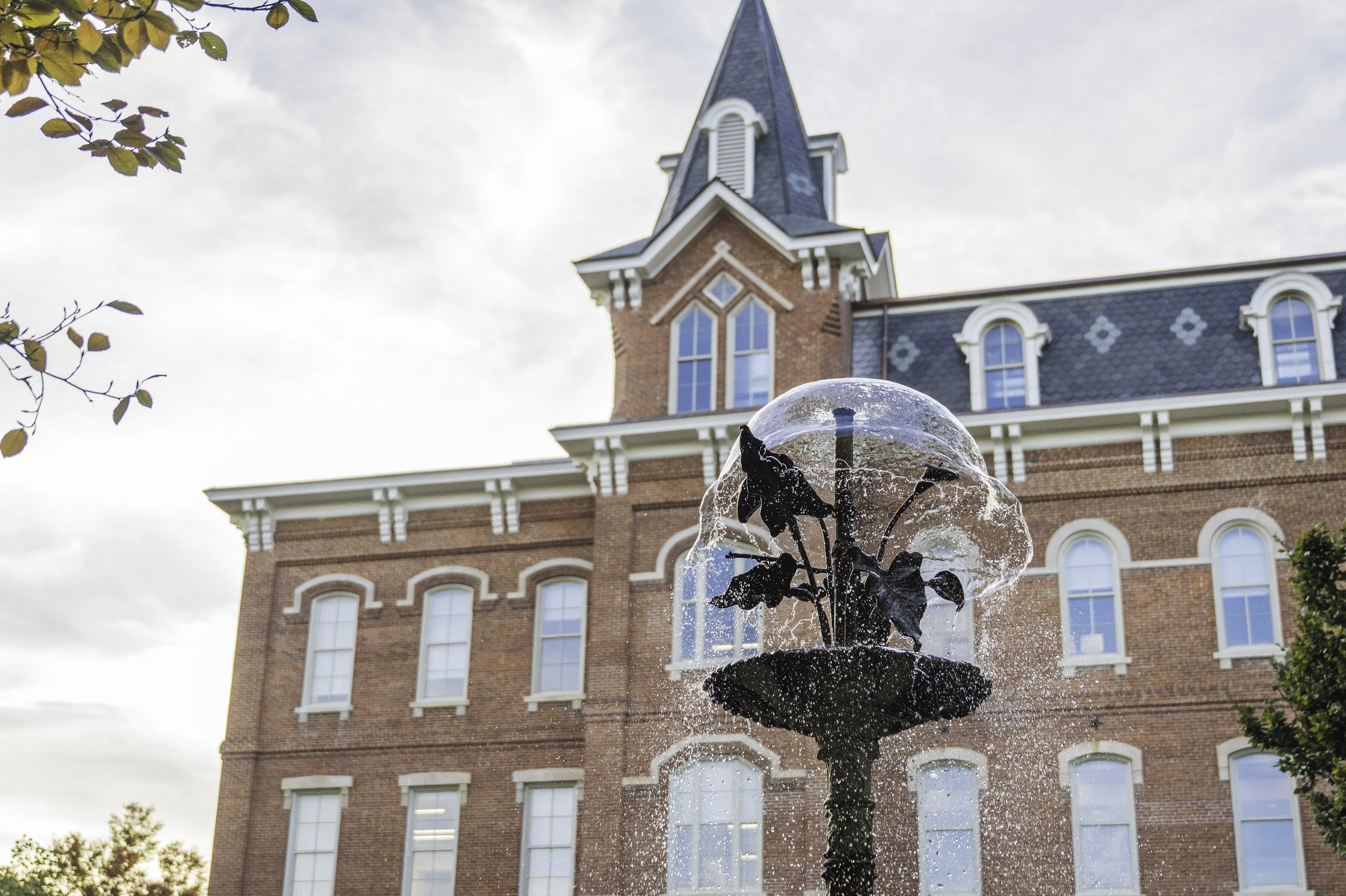 A decorative fountain with a rose sculpture and water spraying outward, in front of a historic brick building with arched windows and a steep roof.