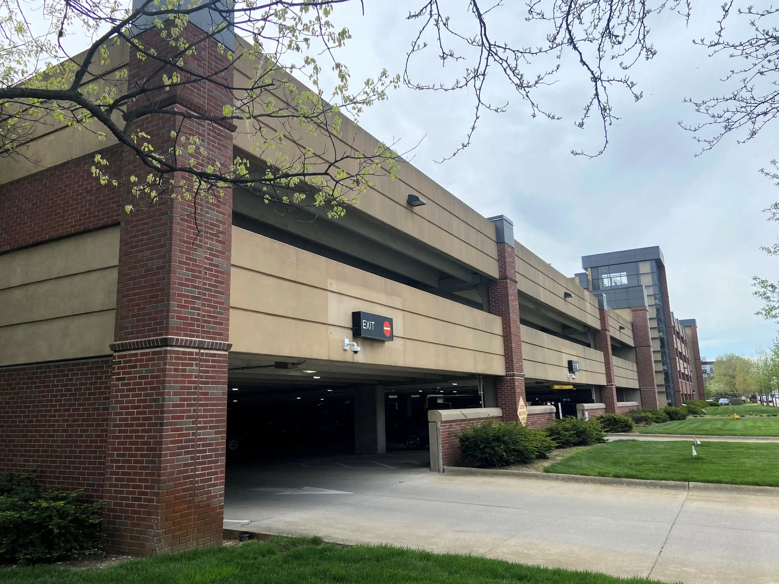 Multi-story parking garage with a brick and concrete exterior, surrounded by green grass and trees, under a partly cloudy sky.