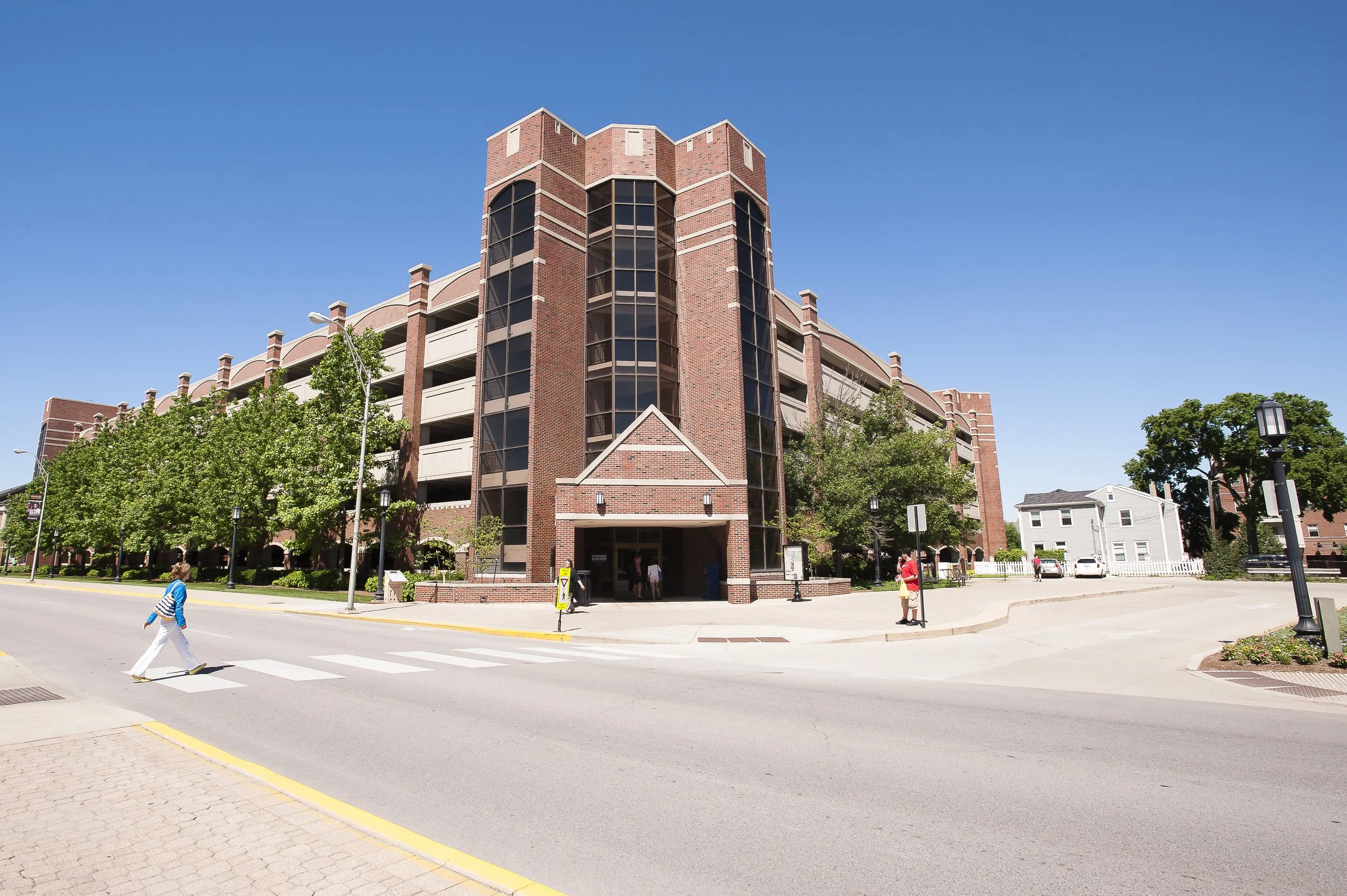 Street view of a multi-story brick parking garage with trees in front and a few people walking nearby under a clear blue sky.