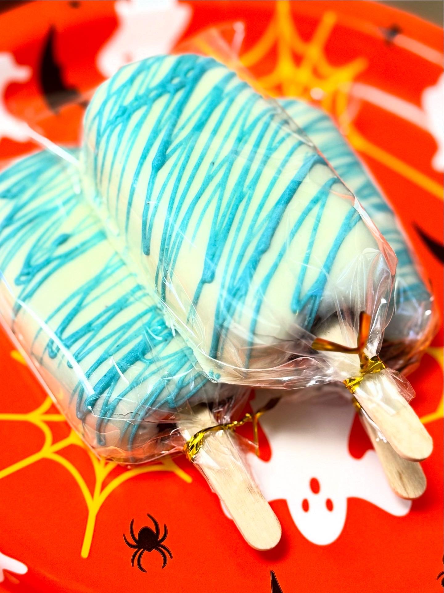 Two Halloween-themed popsicles with blue and white swirl pattern, wrapped in clear plastic and resting on a red plate decorated with orange and black Halloween designs, including a spider and a spider web.
