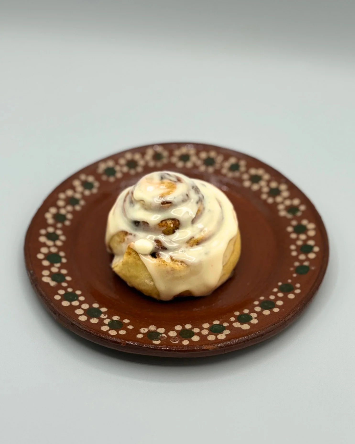 A cinnamon roll with white icing on a small decorative brown plate with green and white dots, against a white background.