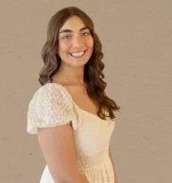 Young woman with long, wavy brown hair wearing a white, short-sleeved dress, smiling against a plain background.