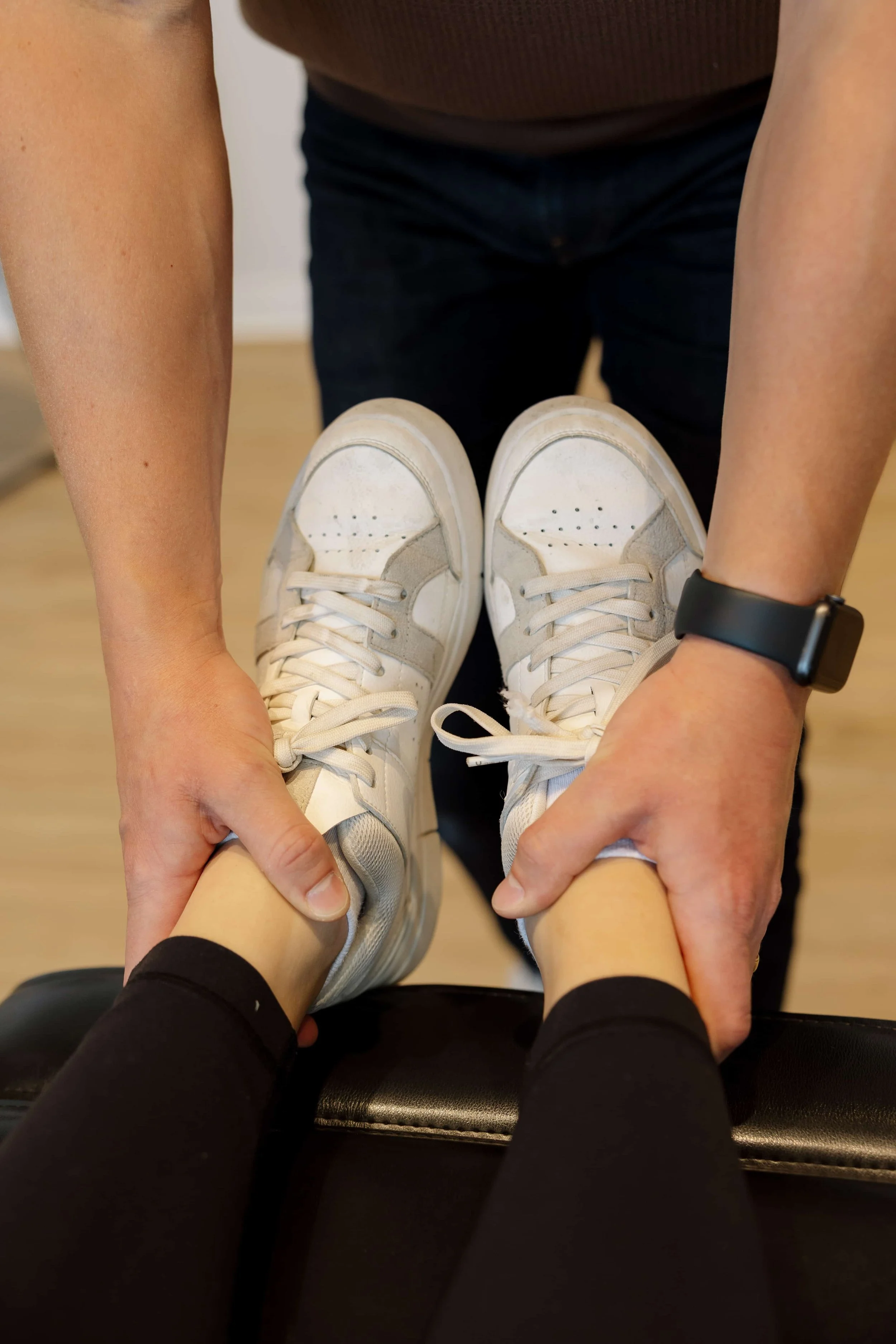 Person receiving physical therapy or chiropractic adjustment, with hands on the ankles and feet, wearing white sneakers, black leggings, a black watch, and a brown top.