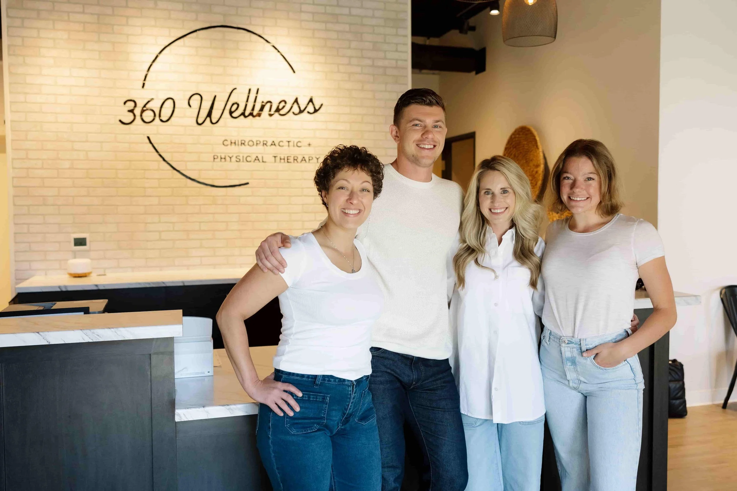 A group of four smiling people standing together inside a wellness center called 360 Wellness. The center offers chiropractic and physical therapy services, with a sign on a brick wall behind them.