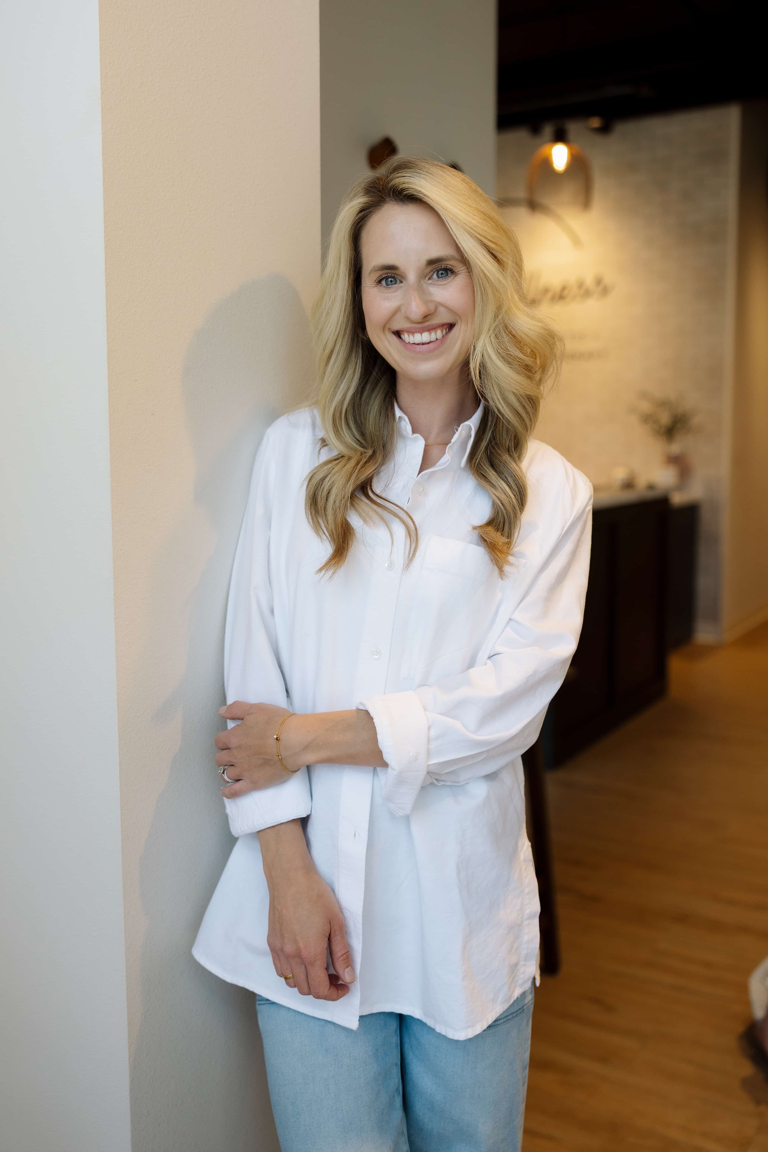 A woman with long, wavy blonde hair and blue eyes smiling while standing in an indoor setting, wearing a white button-up shirt and light blue jeans.