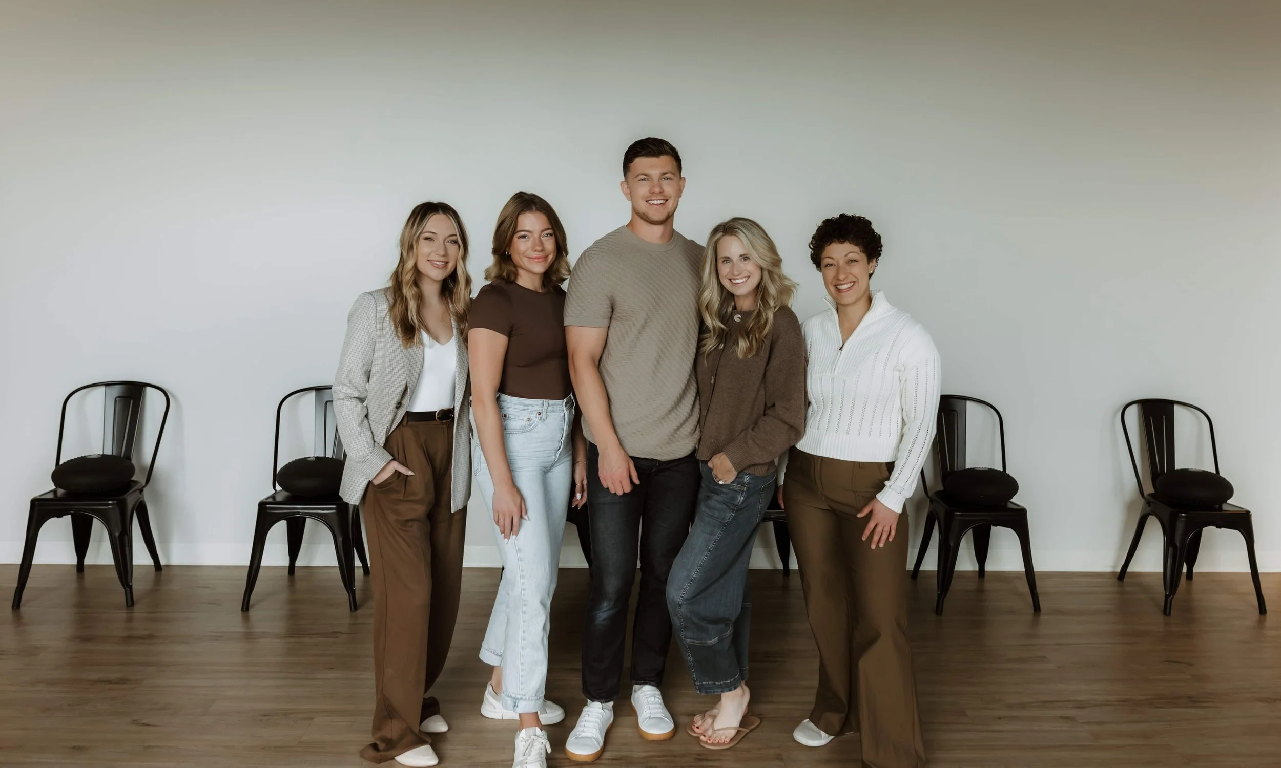 Group of five diverse young adults standing together in a room with a white wall and black chairs behind them, smiling at the camera.