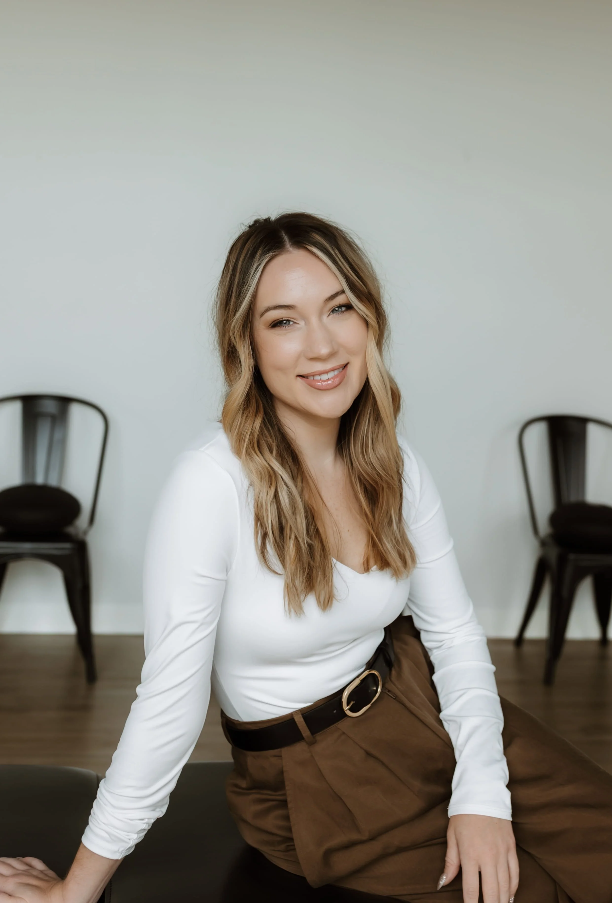 A young woman with wavy blonde hair sitting on a black chair in a minimalistic room with two black chairs in the background, wearing a white long-sleeve top and brown pants.