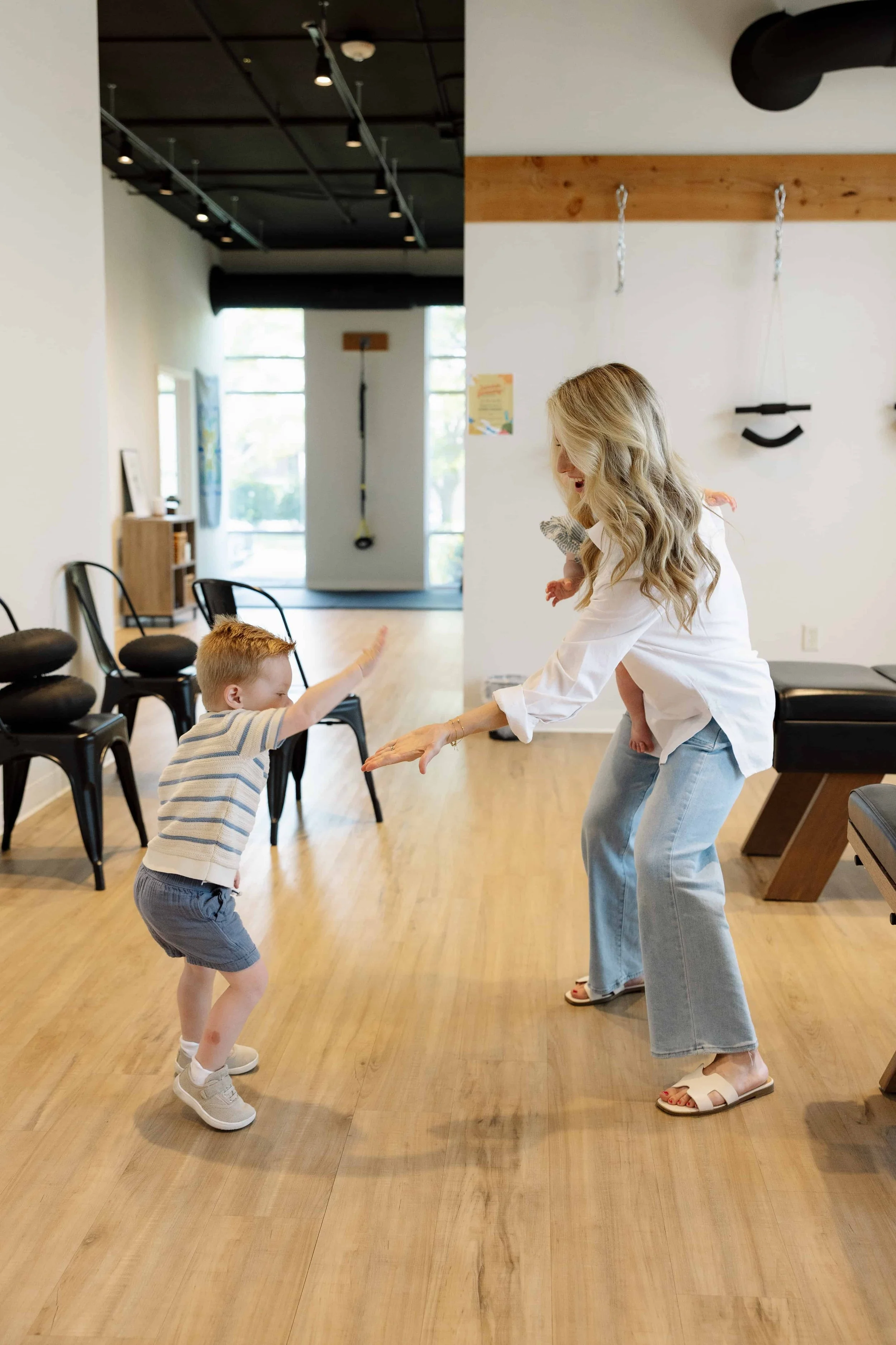 A woman and a young boy are playing a game of high five in a fitness or therapy studio, with a woman holding a baby on her side.