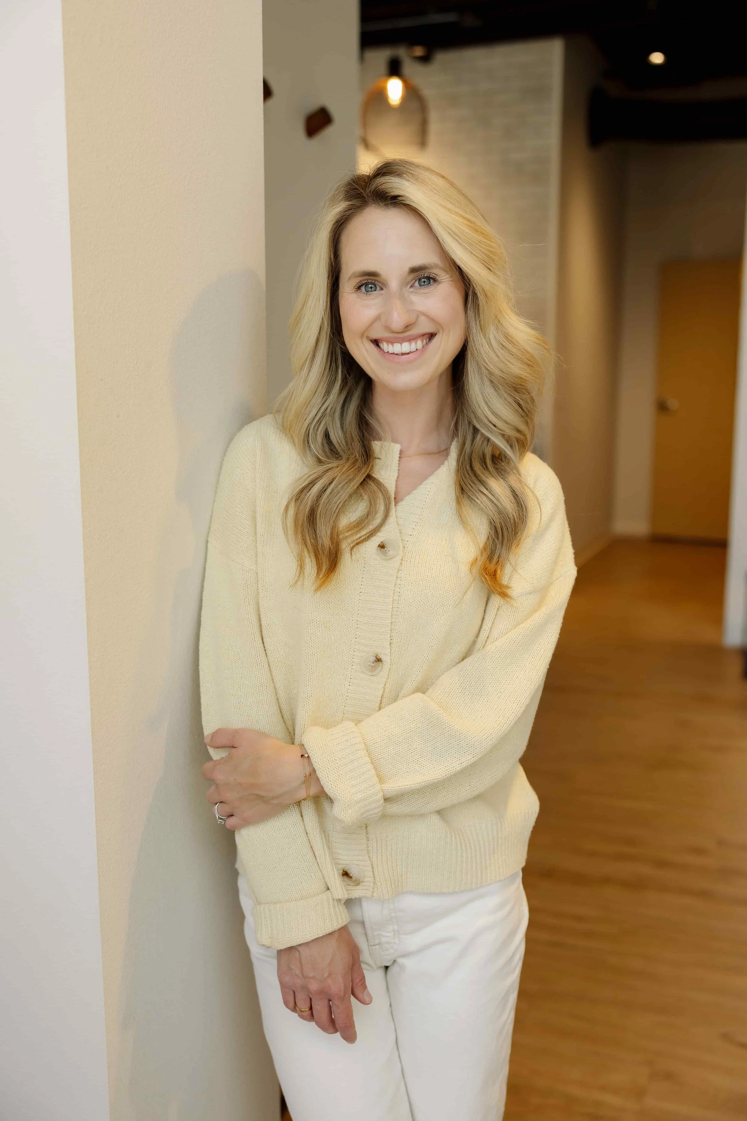 A woman with blonde wavy hair wearing a cream-colored sweater and white pants, smiling and standing against a wall in an indoor setting.