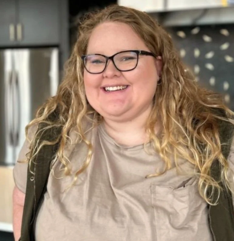 A woman with long curly blond hair, glasses, and a big smile, wearing a beige shirt and a dark vest.