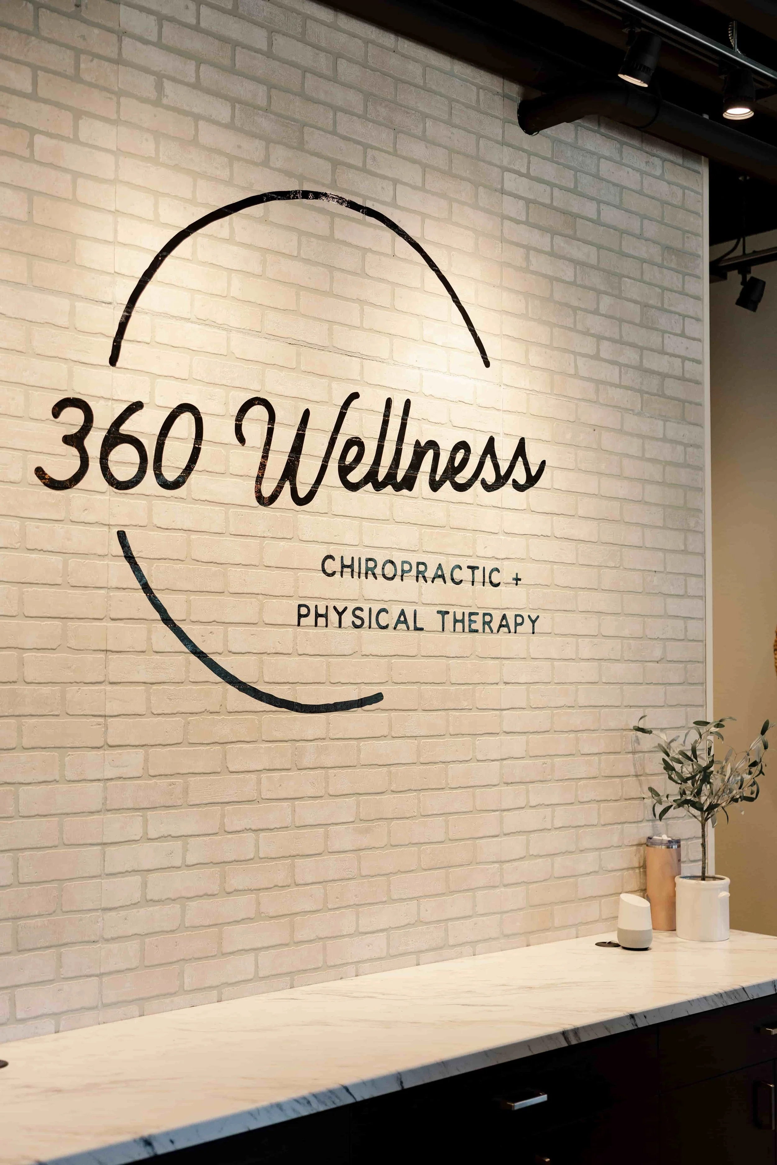Interior wall with the logo of 360 Wellness, which offers chiropractic and physical therapy services, on a white brick wall. There is a white marble counter with a small plant and a white device.