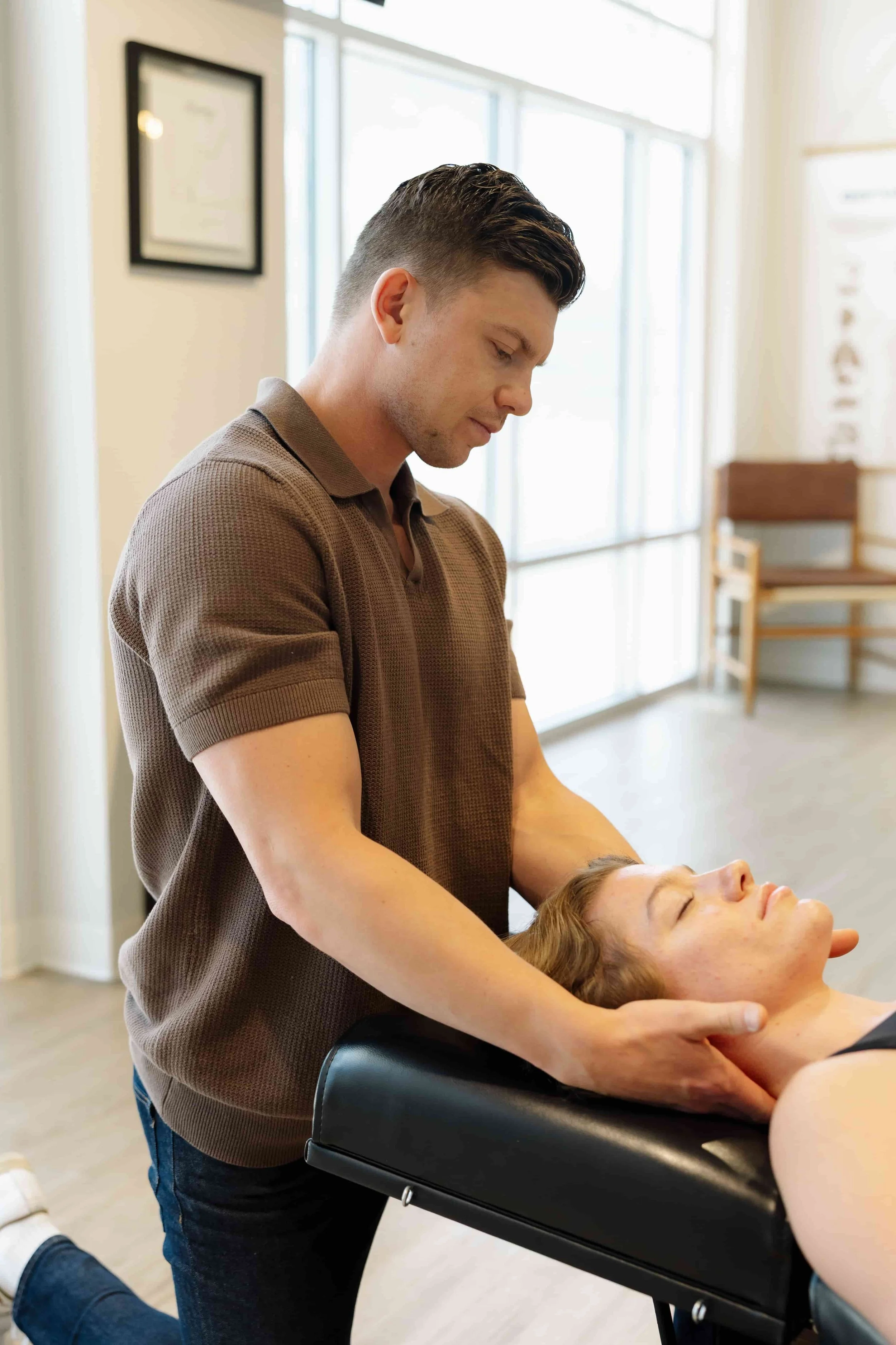 A man receiving chiropractic treatment from a healthcare professional in a clinic with large windows and framed certificates on the wall.