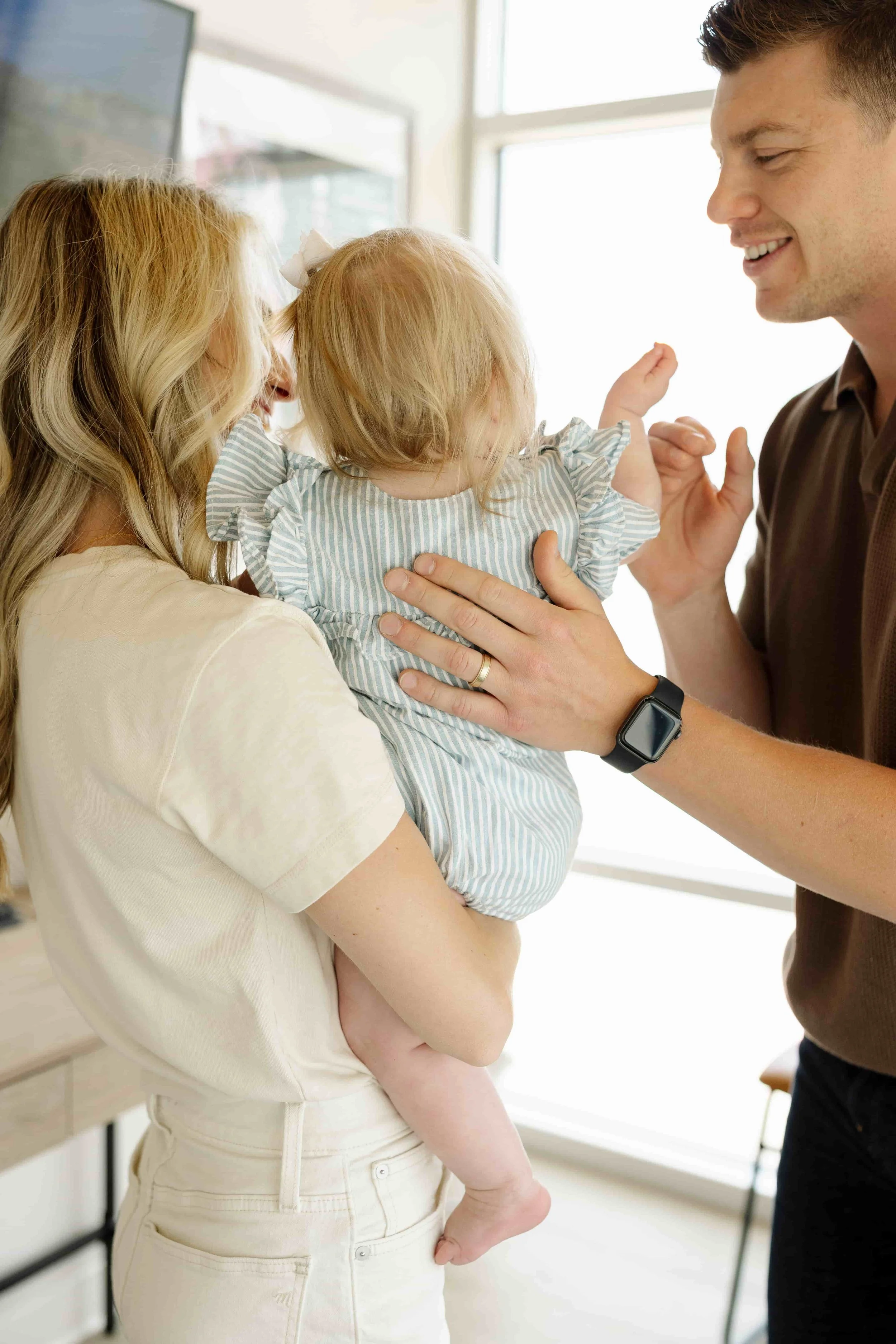 A woman holding a young girl in her arms, while a man smiles at them, touching the girl's hand in a bright room with large windows.