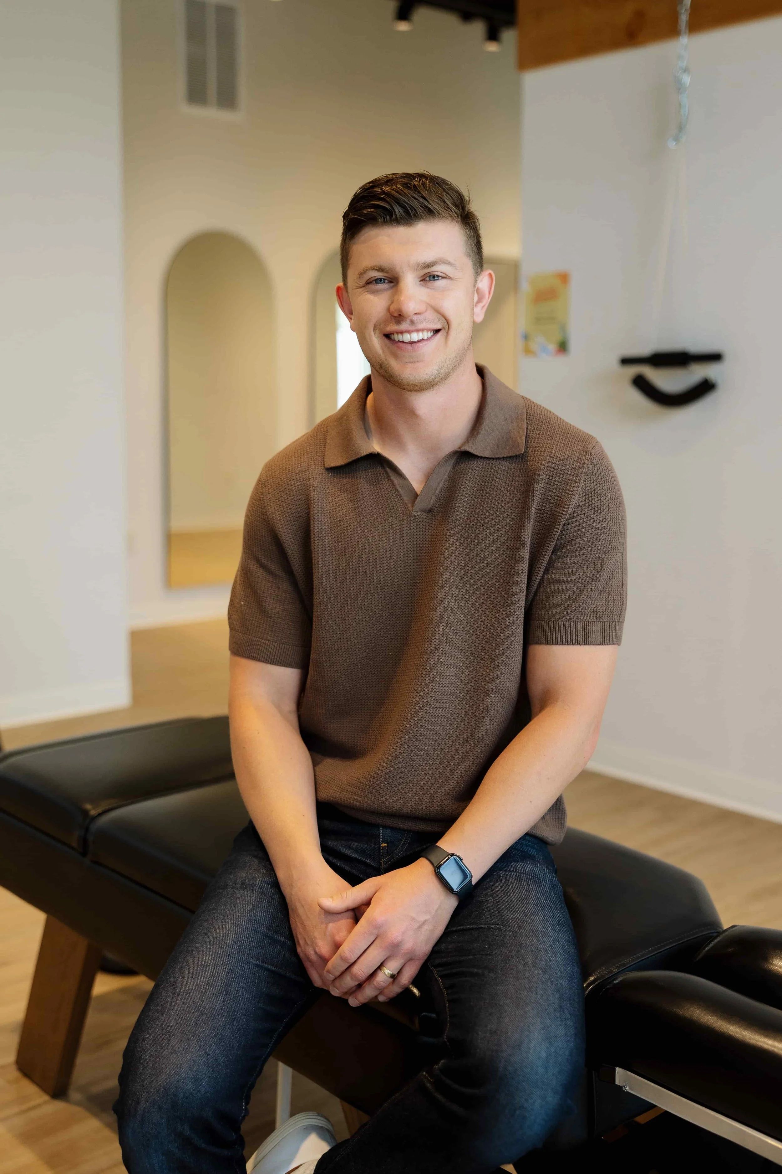 A smiling young man with short brown hair, wearing a brown polo shirt, jeans, and a smartwatch, sitting on a black massage table in a well-lit room.