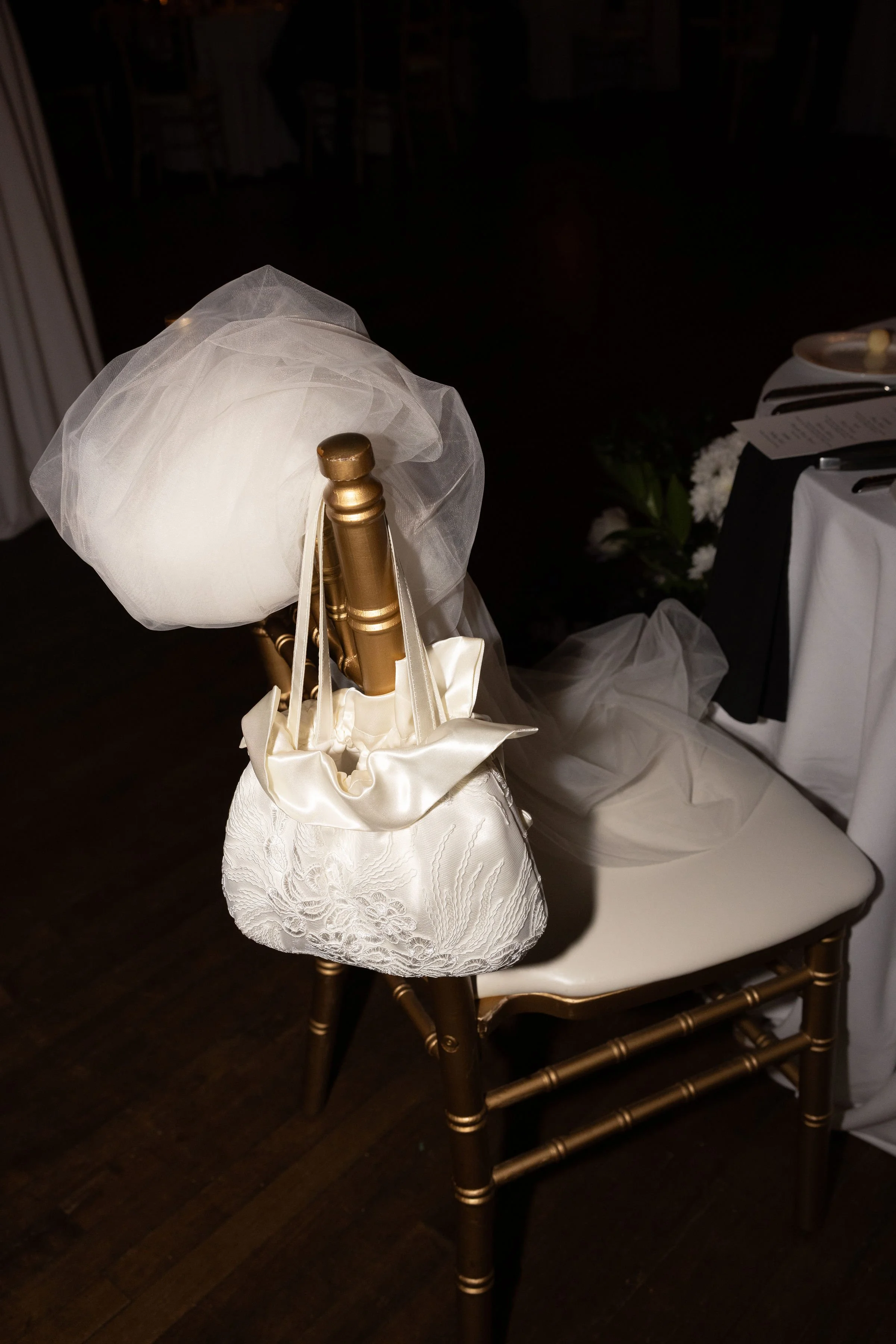 Chair with wedding accessories, including a white veil, a satin handbag, and a small white pillow, at a wedding reception.