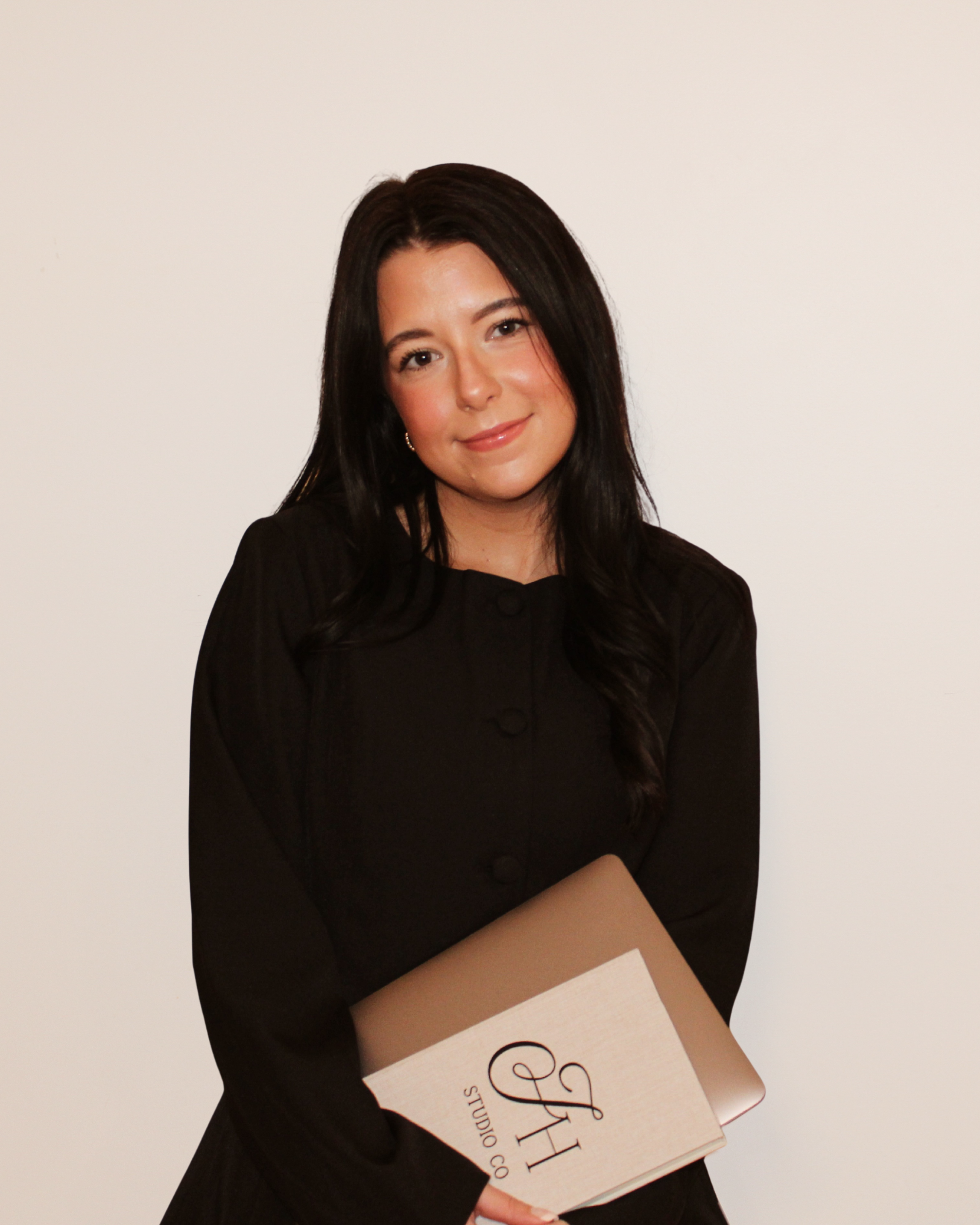 A woman with long dark hair wearing a black top, holding a beige folder with a logo and writing, standing against a plain white background.
