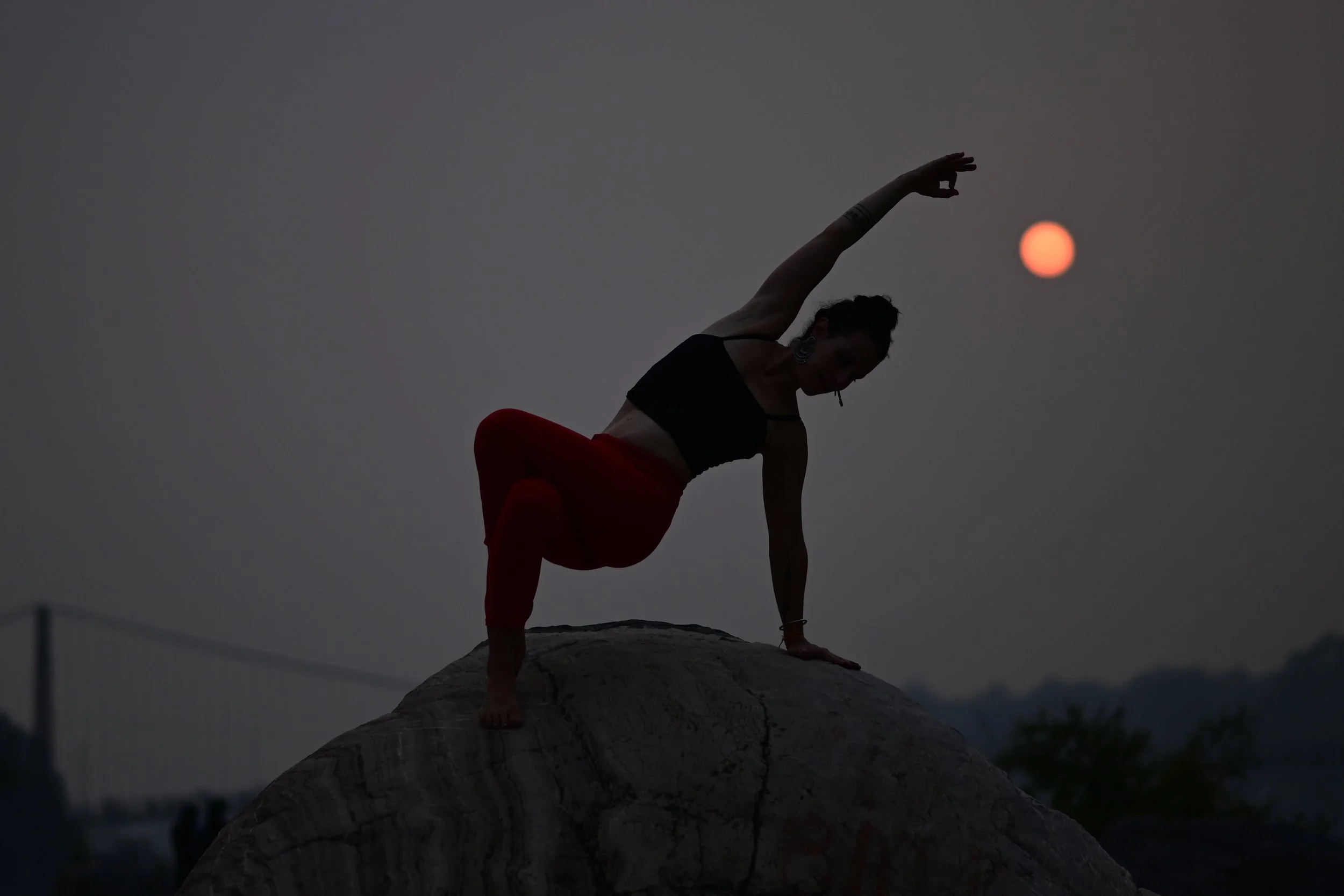 Person practicing yoga on a large rock during sunset, silhouetted against the sky with the sun visible.