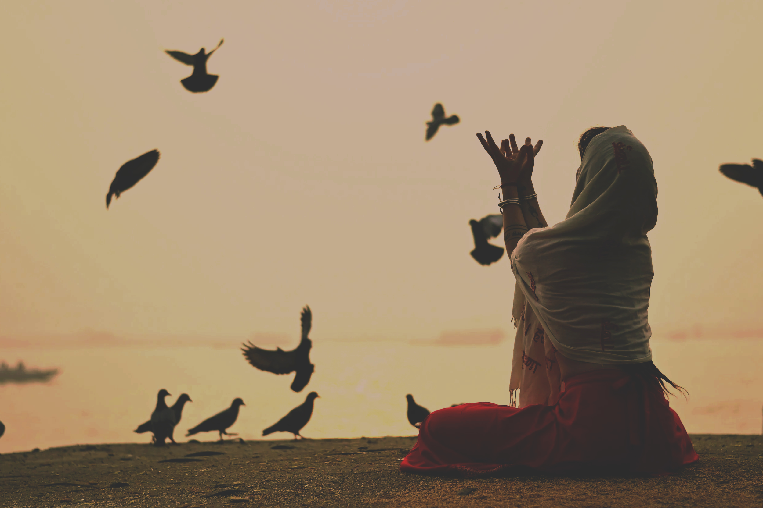 A woman sitting on the ground with her legs crossed, wearing a white headscarf and red skirt, releasing birds into the sky during sunset by a body of water.