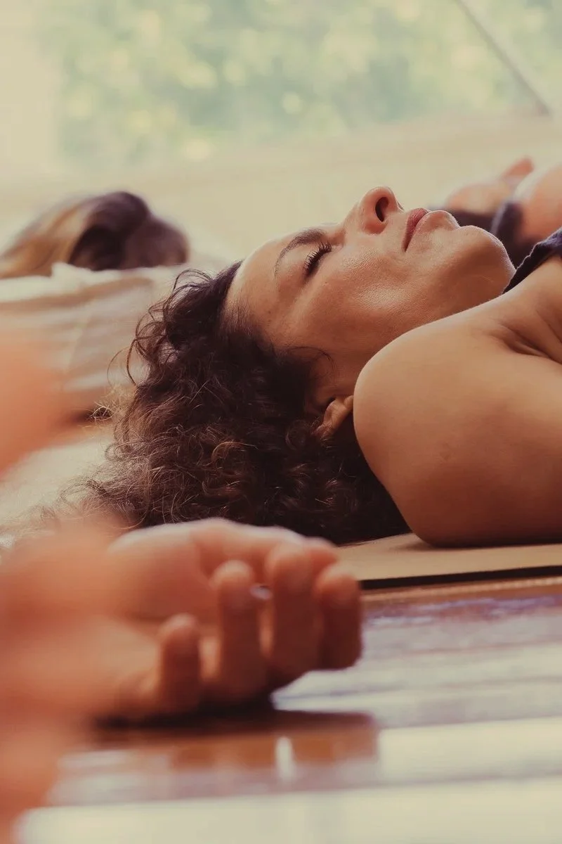 Two people lying on the floor with their eyes closed, possibly meditating or resting, in a bright room with a window and green scenery outside.