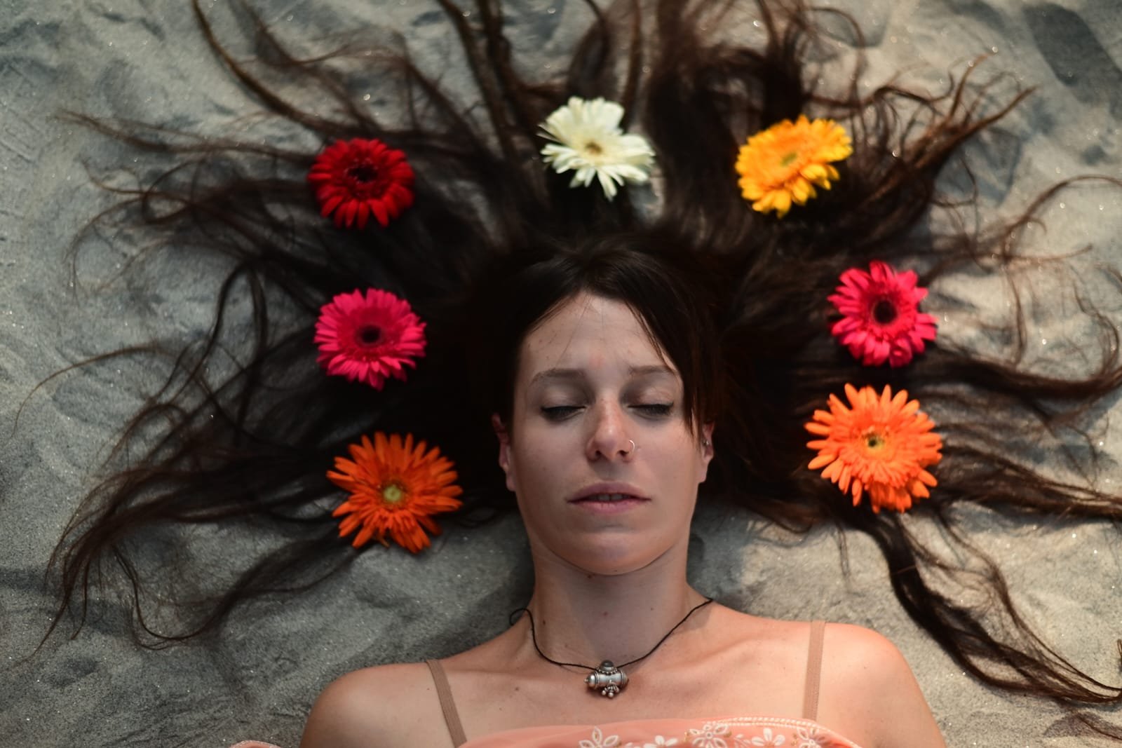 A woman with long dark hair lying on the sand with her eyes closed. Her hair is decorated with colorful flowers, including orange, pink, red, white, and yellow. She wears a thin strappy top and a necklace with a small decorative charm.