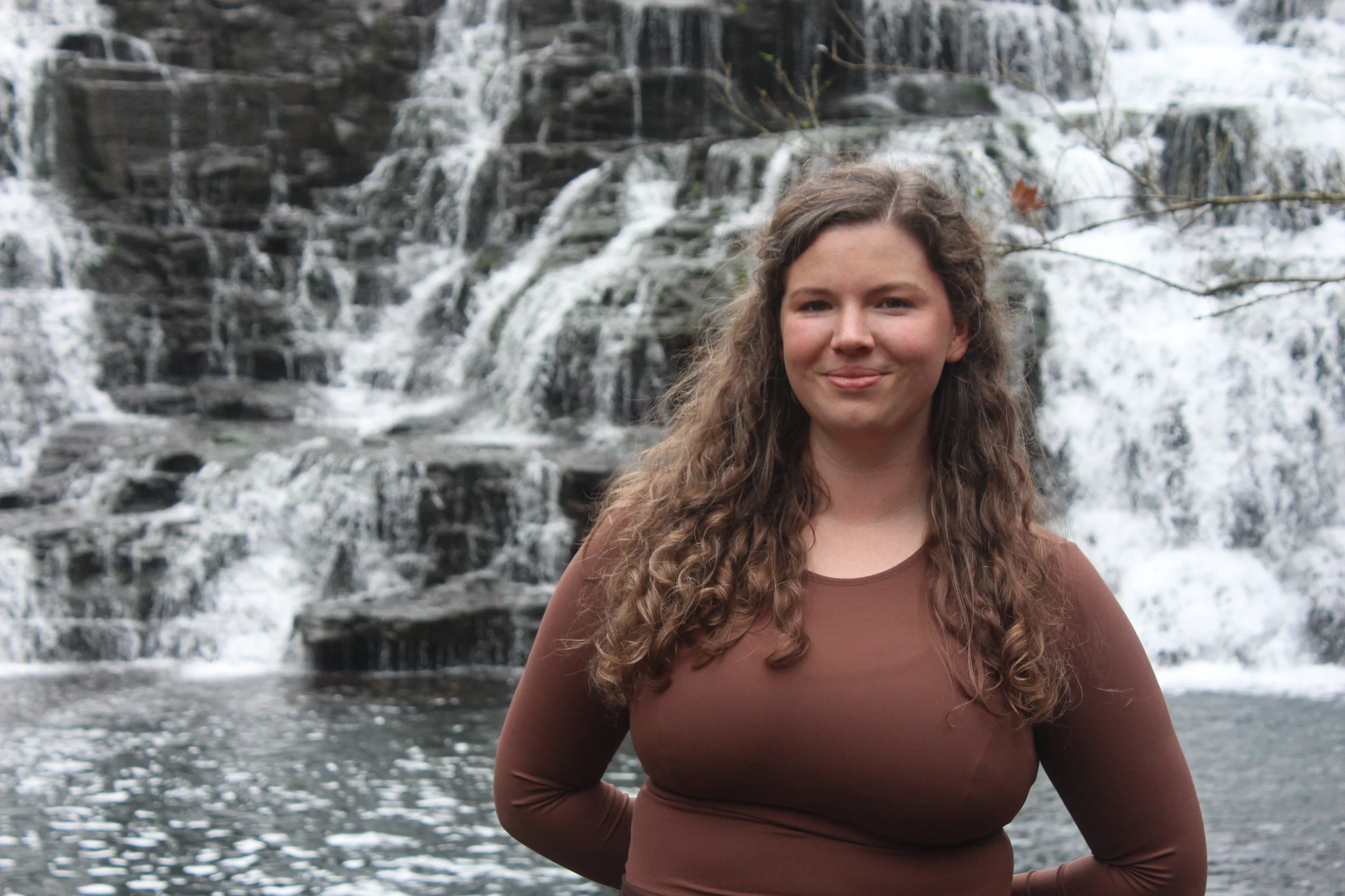 A young woman with curly hair standing in front of a waterfall during the daytime.